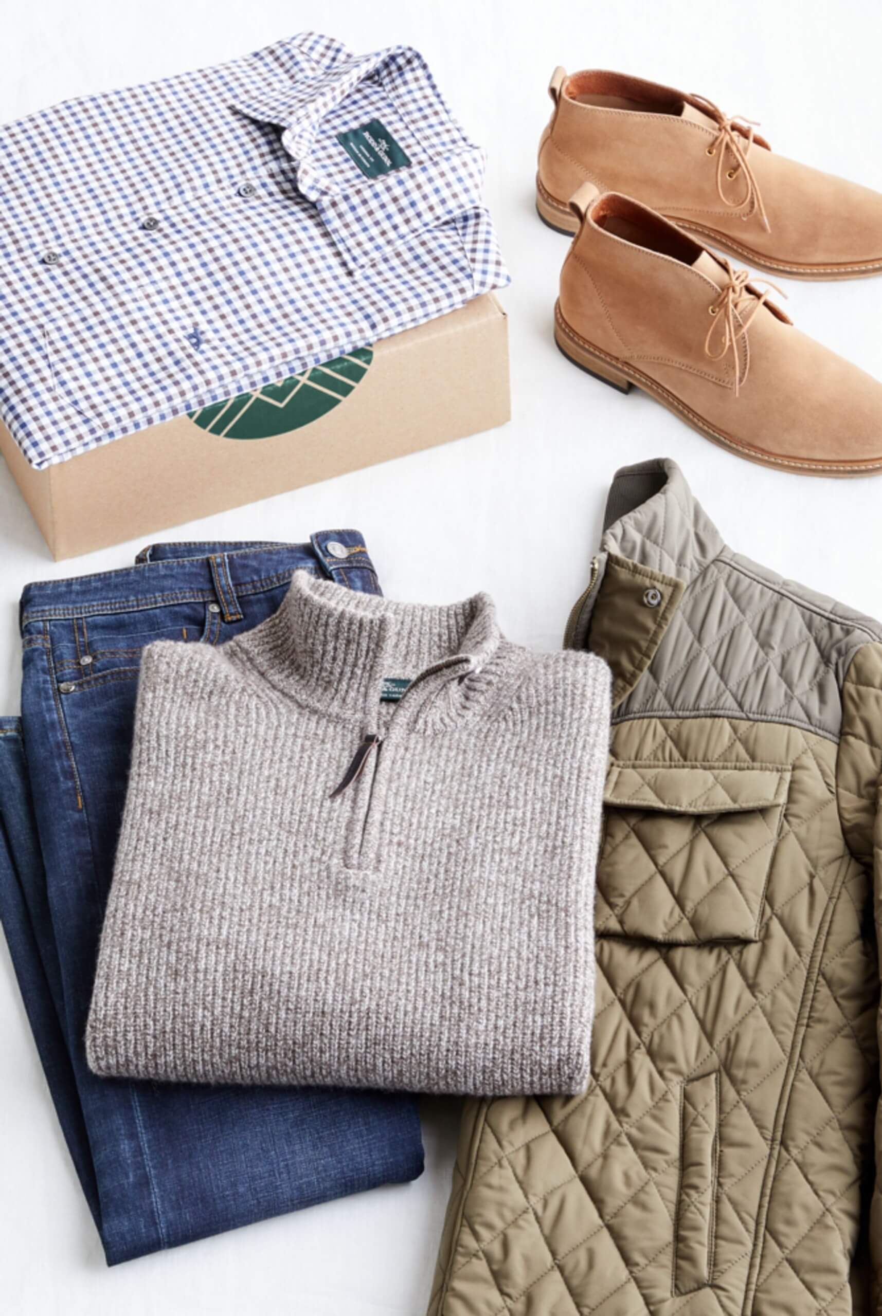 Stitch Fix Men’s outfit laydown featuring a quarter-zip sweater over dark wash jeans and quilted jacket, next to tan chukkas and checkered woven shirt sitting on a Stitch Fix delivery box.