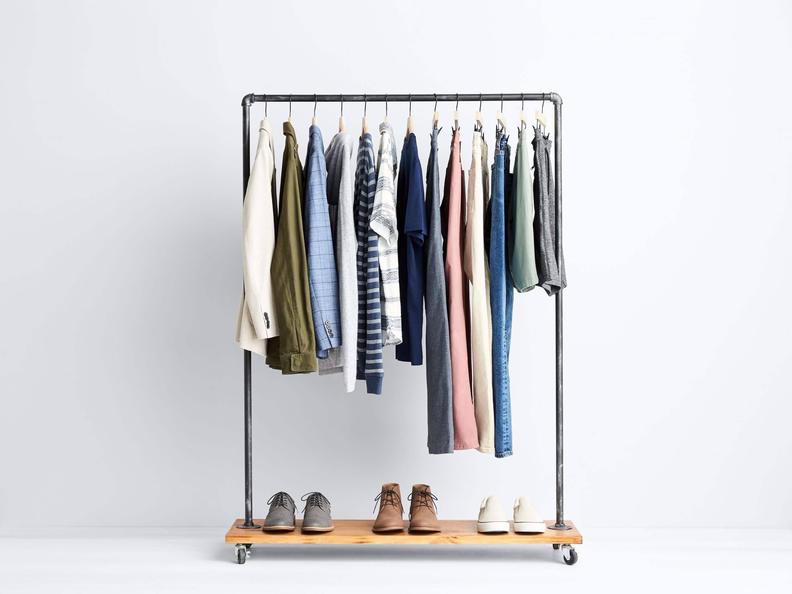 Stitch Fix Men’s rack image featuring beige blazer, green military jacket, blue button-down shirt, grey hoodie, striped shirts, navy polo, grey pants, pink pants, khaki pants, blue jeans, green shorts and grey shorts hanging above grey sneakers, brown chukkas and white sneakers. 