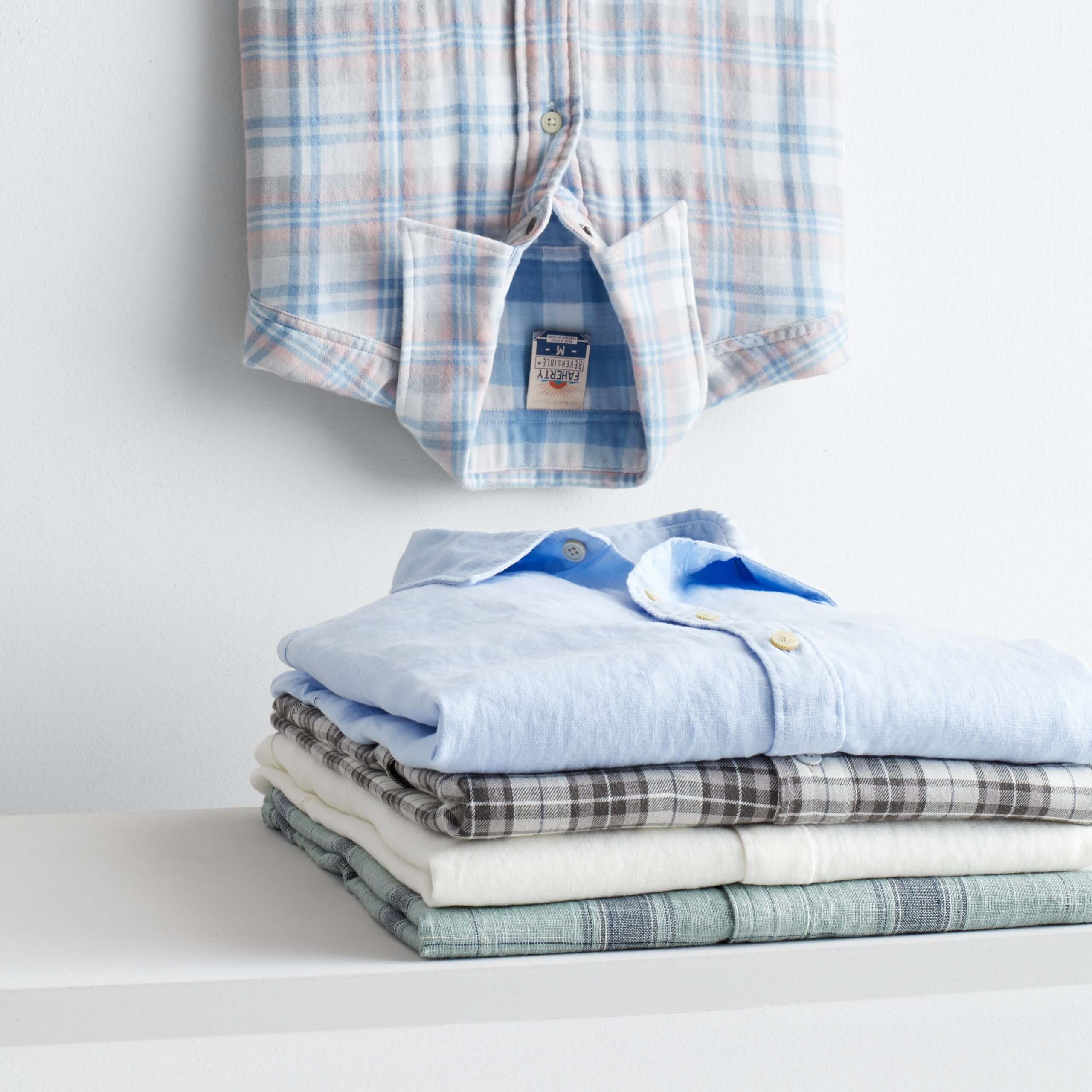 Stitch Fix Men’s folded stack of button-down shirts in various shades of blue, cream and grey and one blue plaid shirt hanging upside down. 