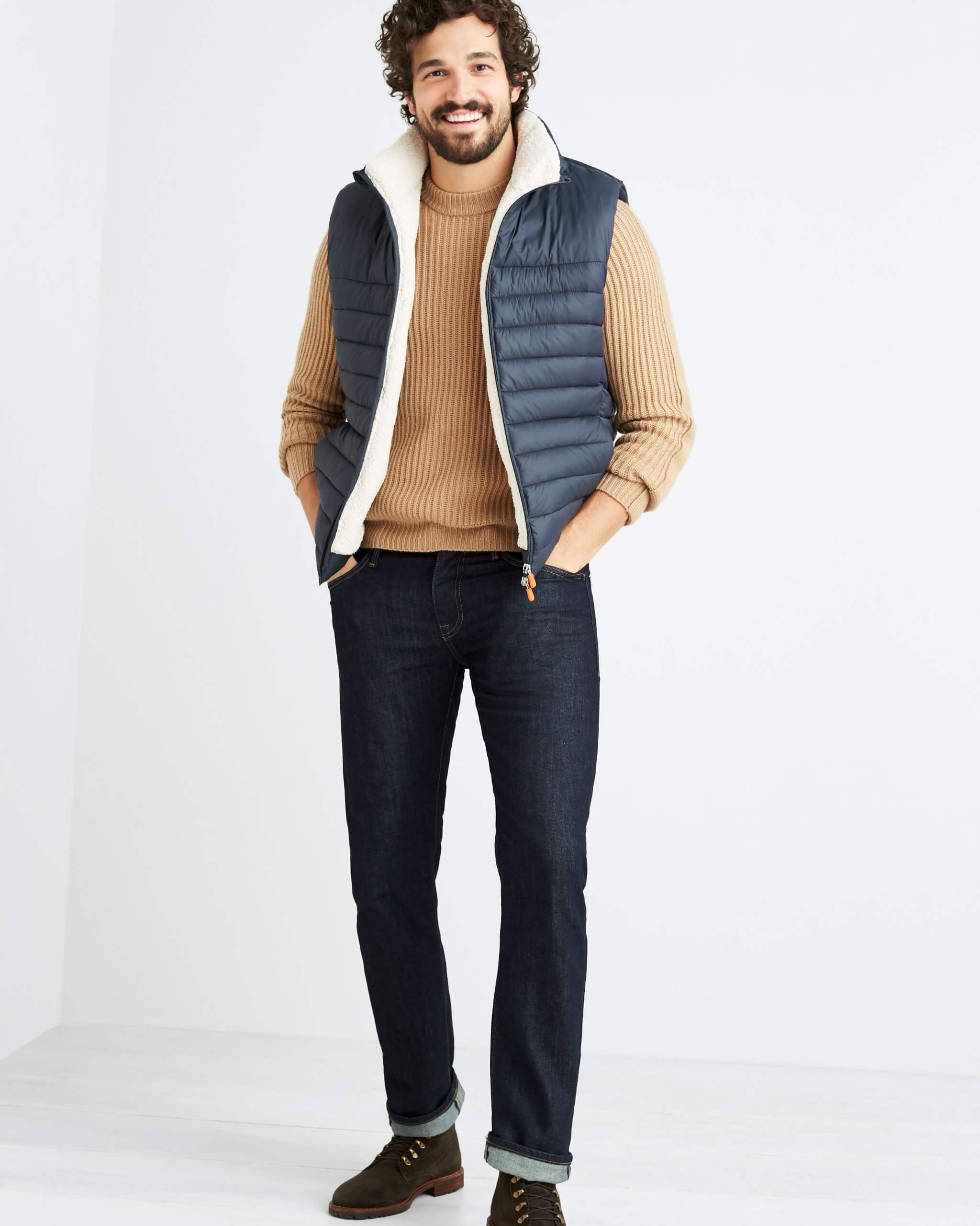 Stitch Fix Men’s model wearing dark wash jeans, brown crewneck sweater, navy puffer vest and brown boots. 