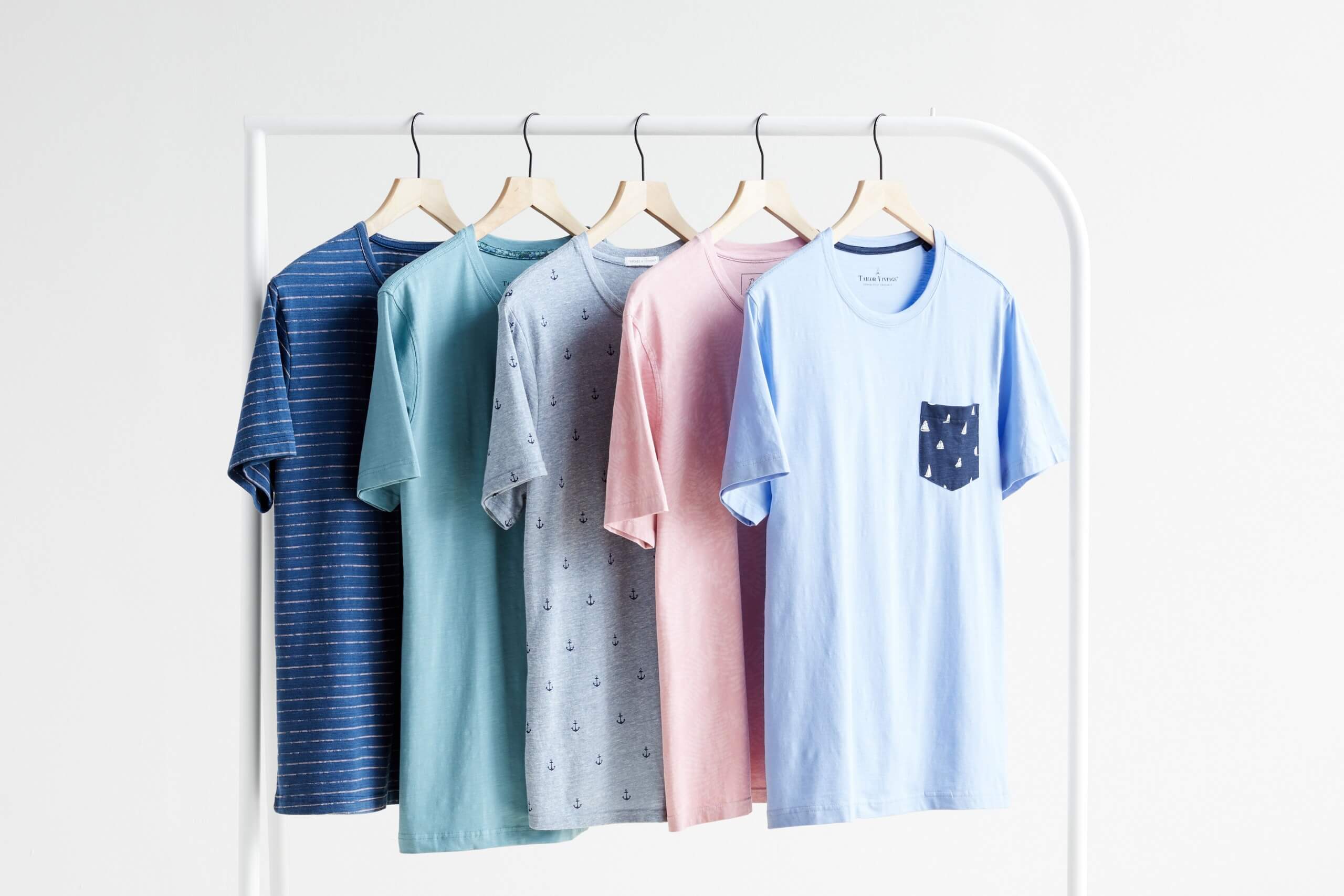 Stitch Fix Men’s baby blue crew neck t-shirt, pink crew neck t-shirt, baby blue anchor print crew neck t-shirt, teal crew neck t-shirt and blue striped crew neck t-shirt hanging on white rack. 