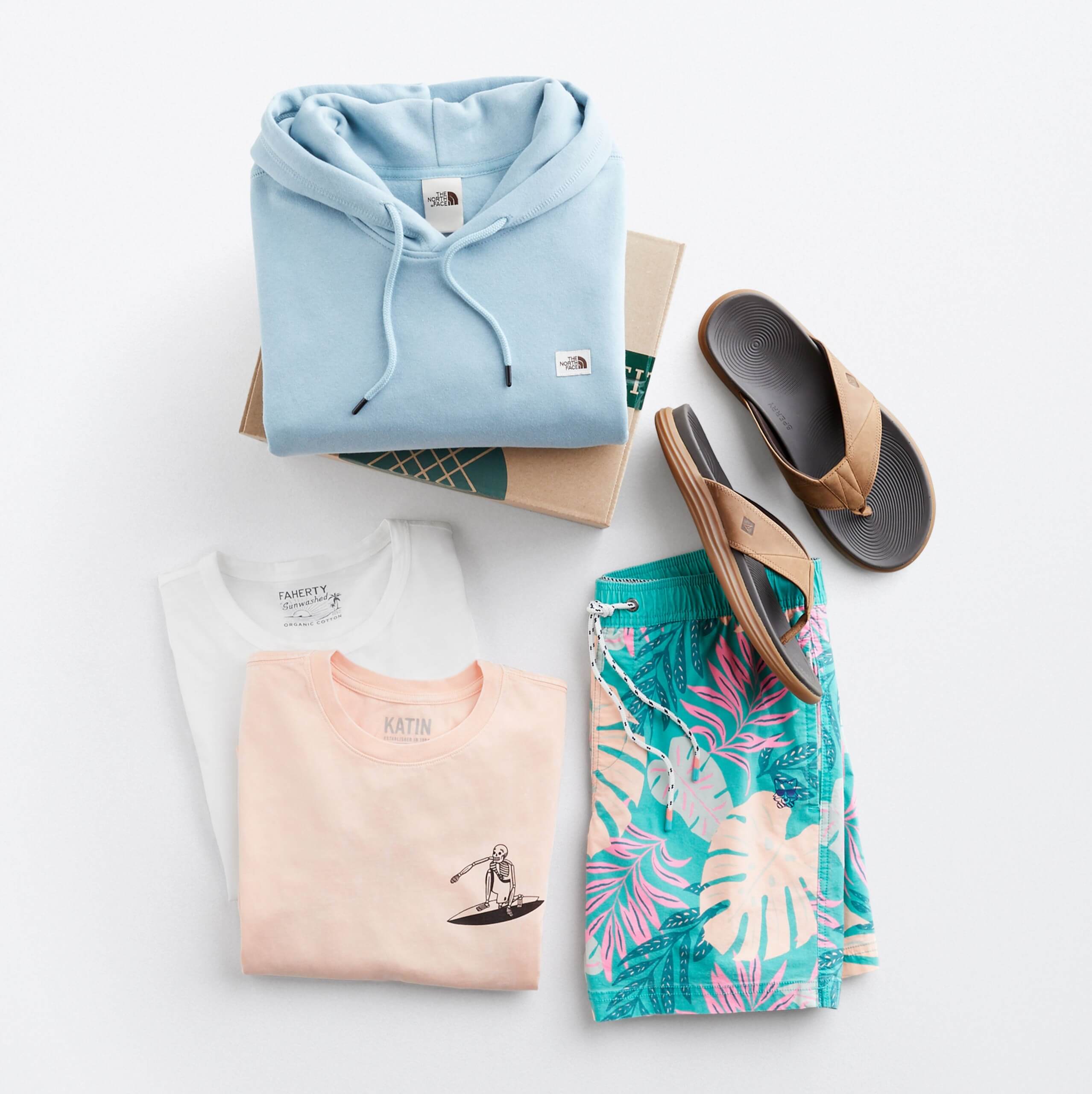 Stitch Fix Men’s outfit laydown featuring a light blue hoodie on a Stitch Fix delivery box, next to tan flip flop sandals, tropical print shorts and two crew neck t-shirts in pink and white. 