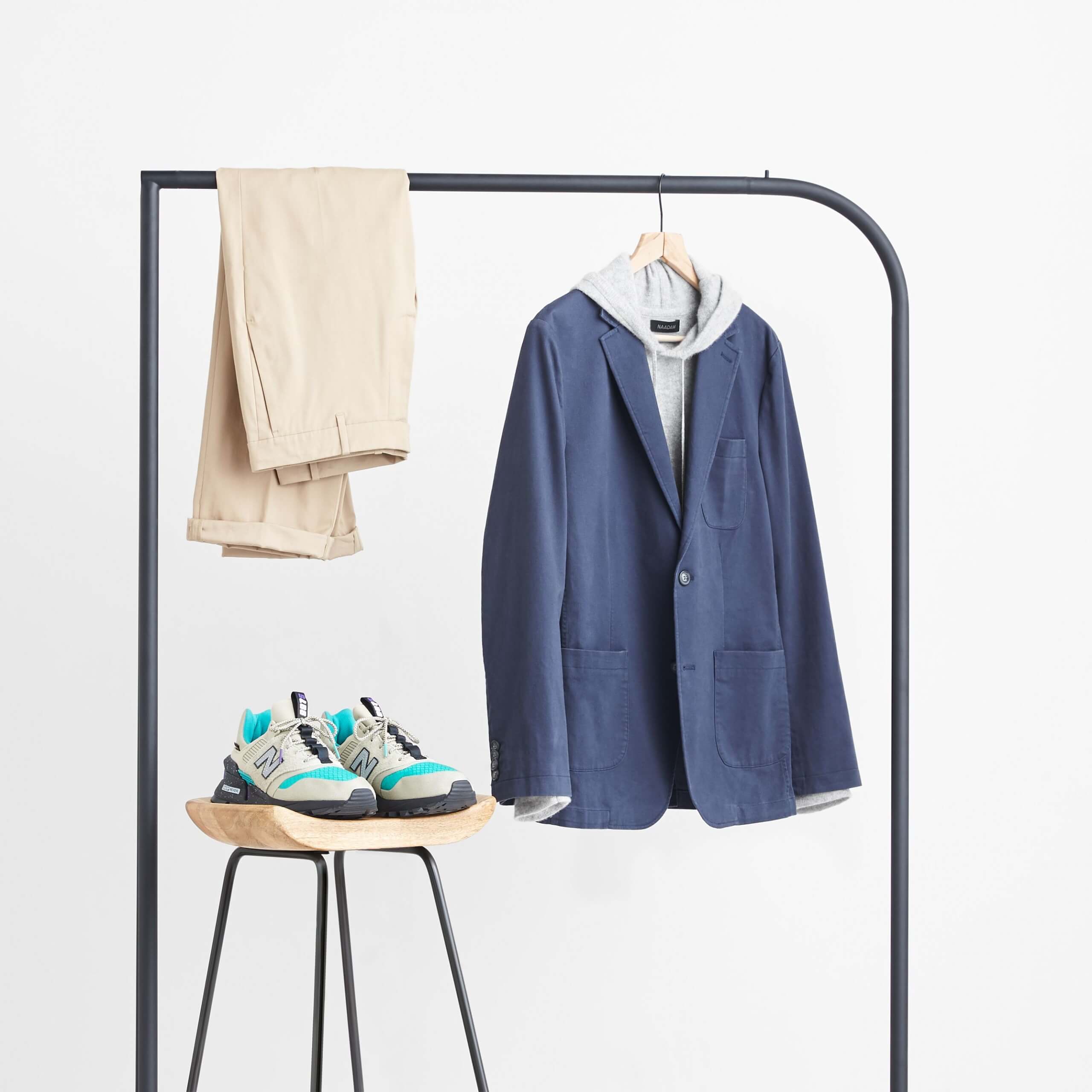 Stitch Fix Men’s navy blazer, grey hoodie and tan chinos hanging on a black rack next to tan nylon sneakers sitting on a wooden stool. 