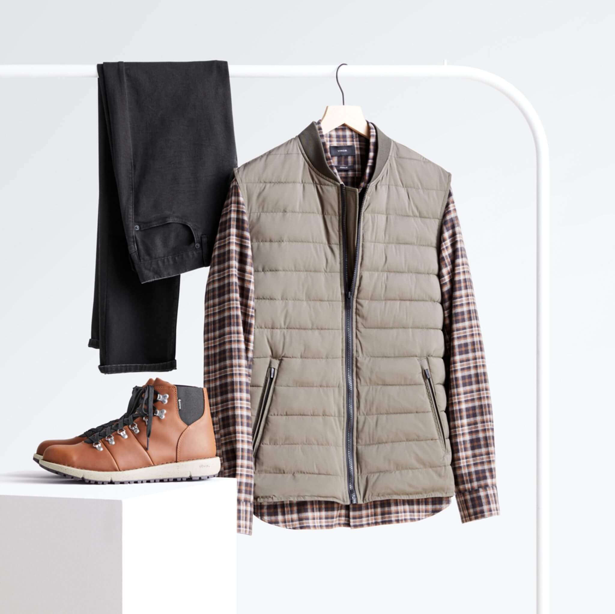 Stitch Fix Men’s black slim-fit jeans, long-sleeved woven plaid button-down shirt and bomber vest hanging on a white rack with brown leather boots on a white podium. 