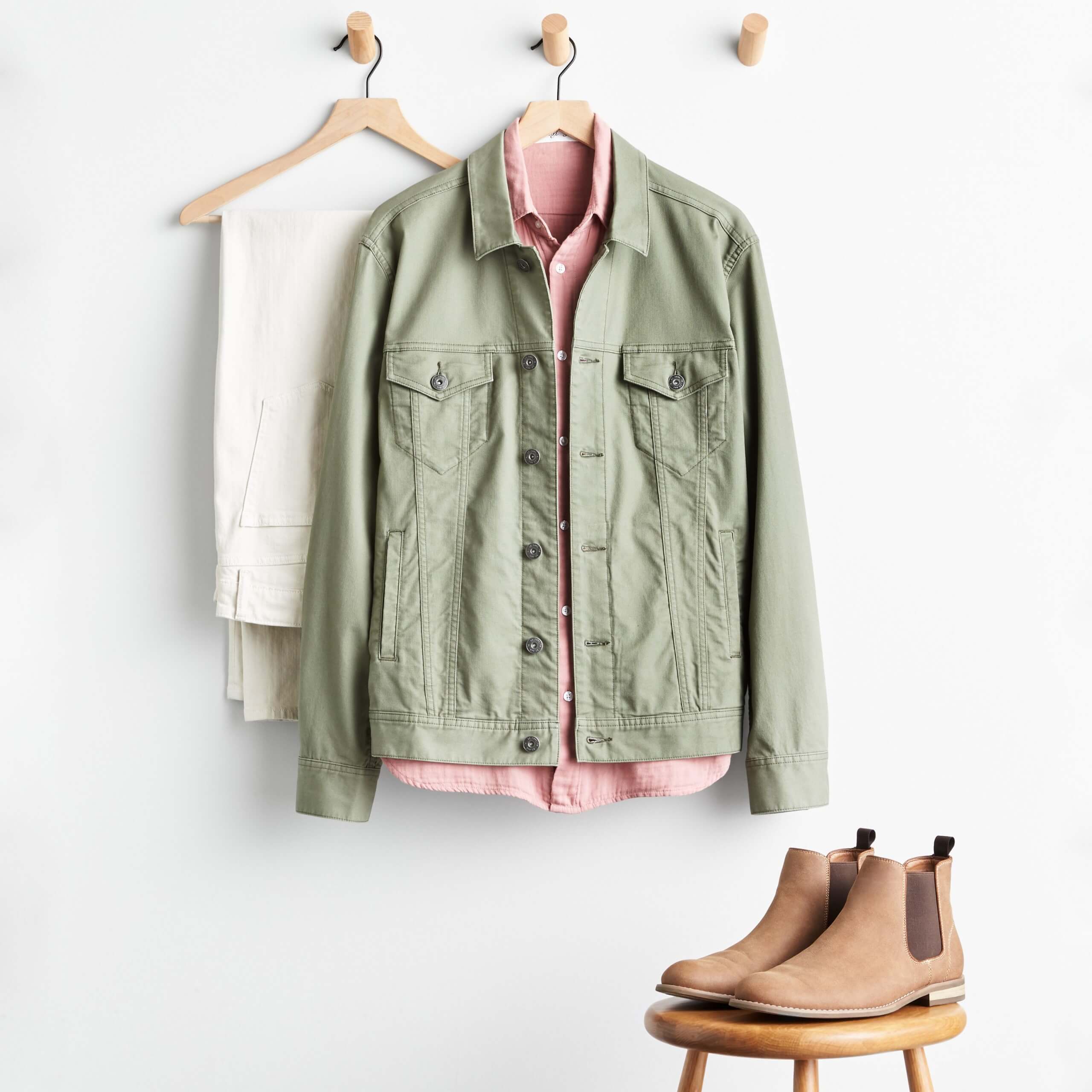 Stitch Fix Men’s khaki pants hanging on wooden hanger, olive denim jacket over pink woven shirt on wooden hanger and tan chelsea boots on a wooden stool. 