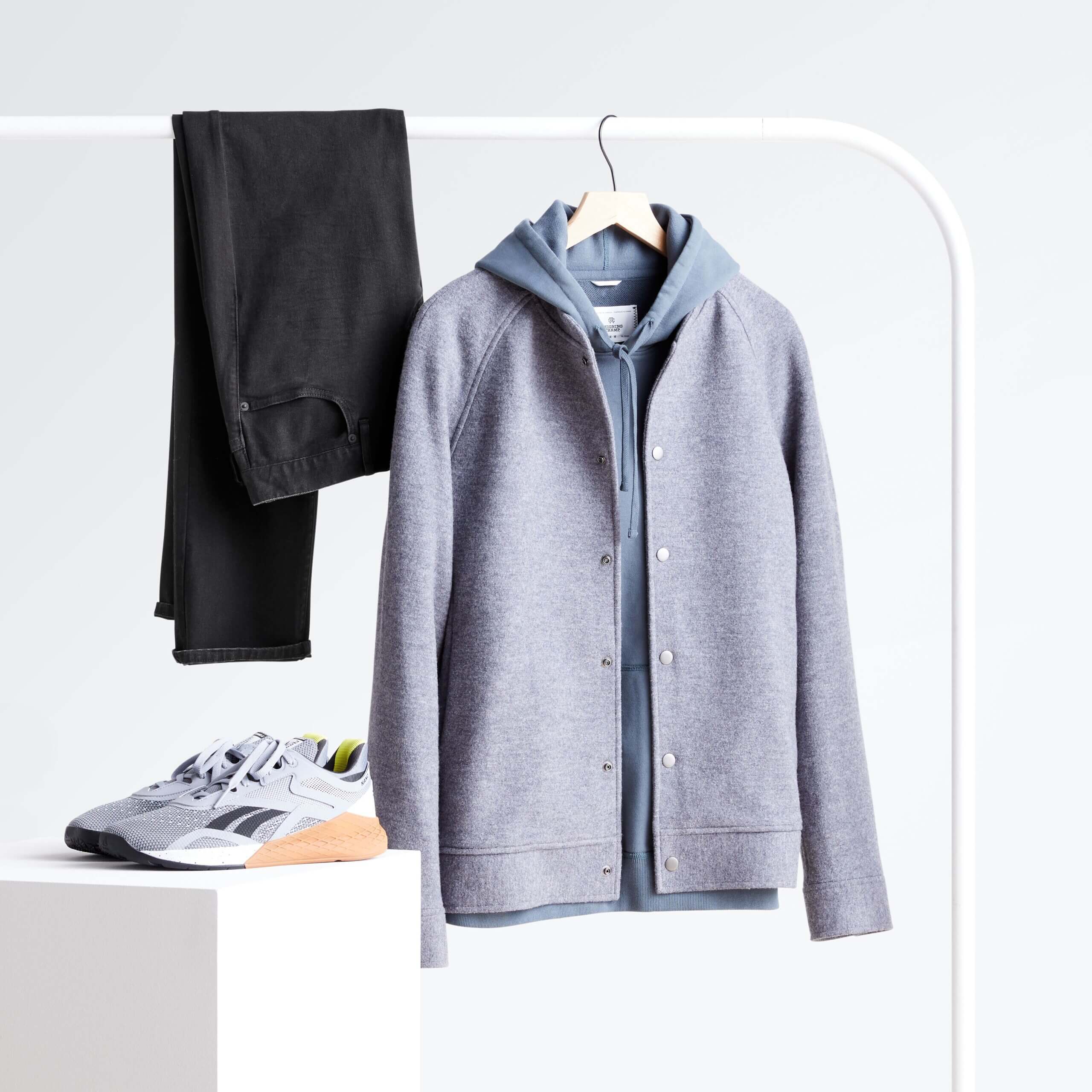 Stitch Fix men's rack image featuring grey wool bomber over blue hoodie and black jeans hanging on white rack next to gray active sneakers on white block.