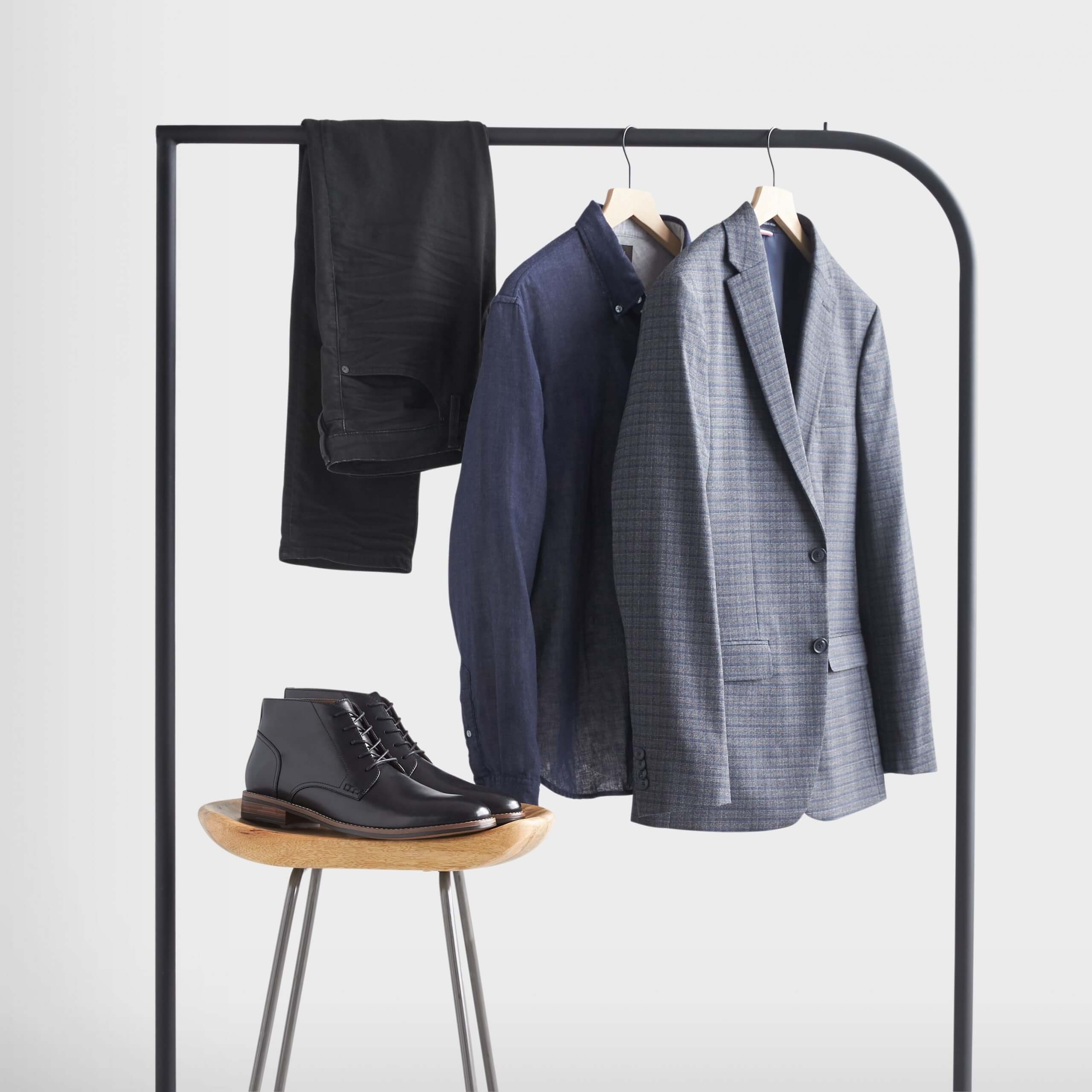 Stitch Fix Men's rack image featuring blue blazer, navy blue dress shirt and black jeans hanging on black rack, next to black leather chukkas on wooden stool. 