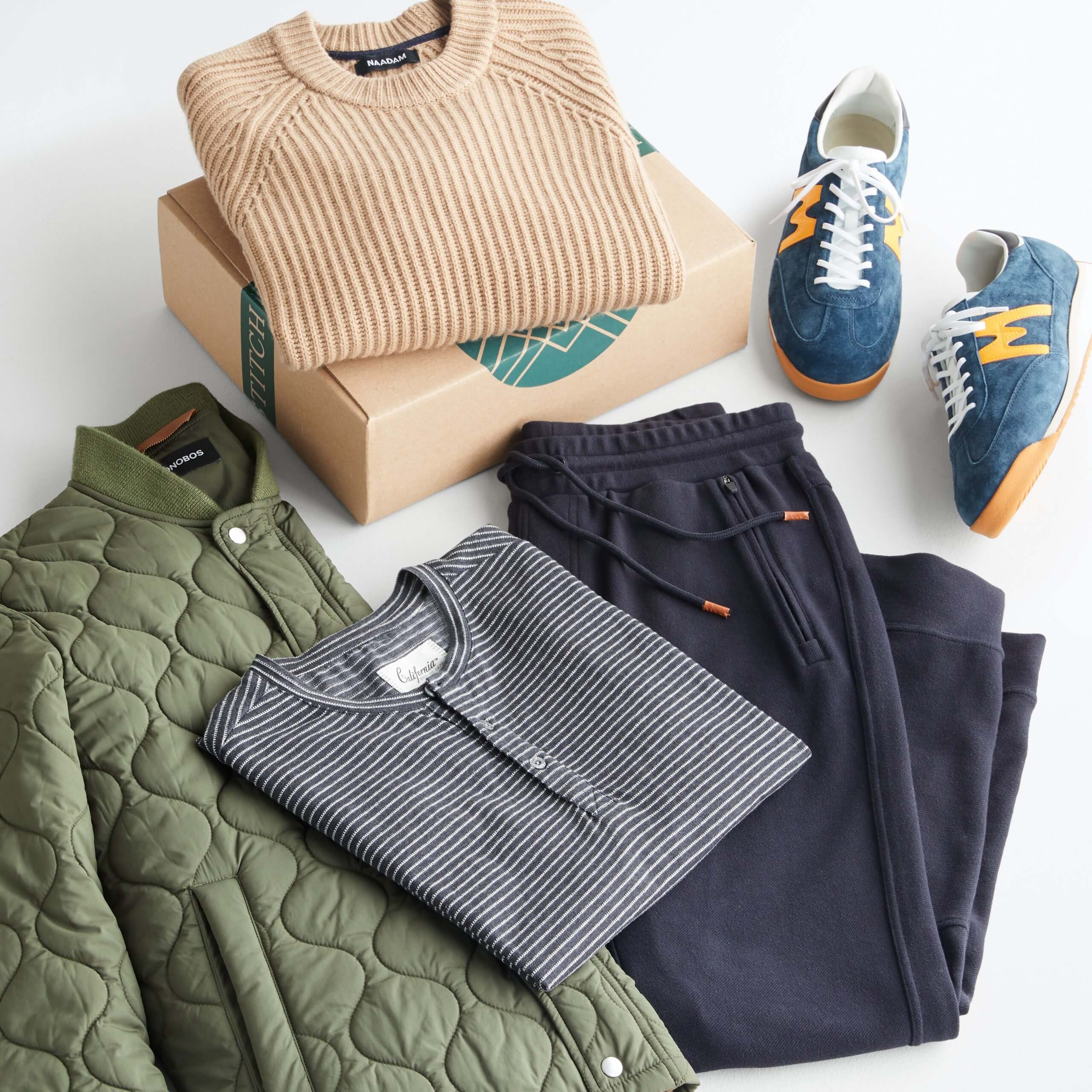 Stitch Fix Men’s outfit laydown featuring camel crewneck sweater folded on Stitch Fix next to blue retro suede sneakers, blue joggers, striped henley and olive quilted bomber jacket.