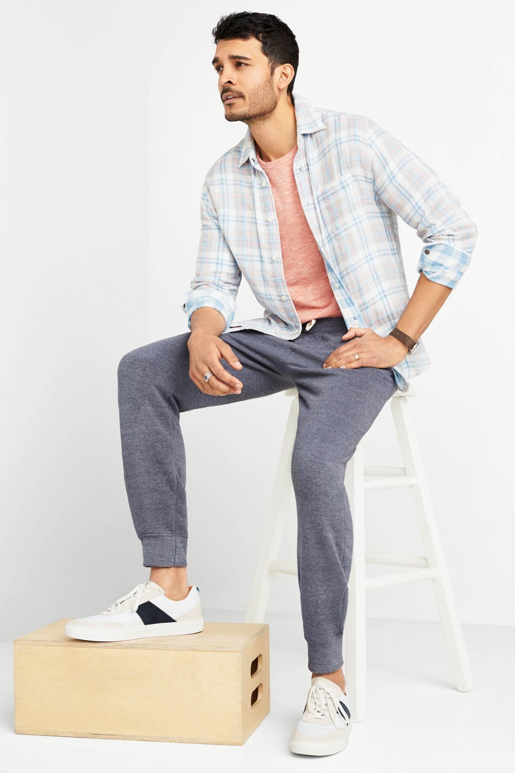 Stitch Fix Men’s model sitting on a white stool wearing pink terry crew sweatshirt under a plaid button-down shirt with fleece pants and white sneakers. 