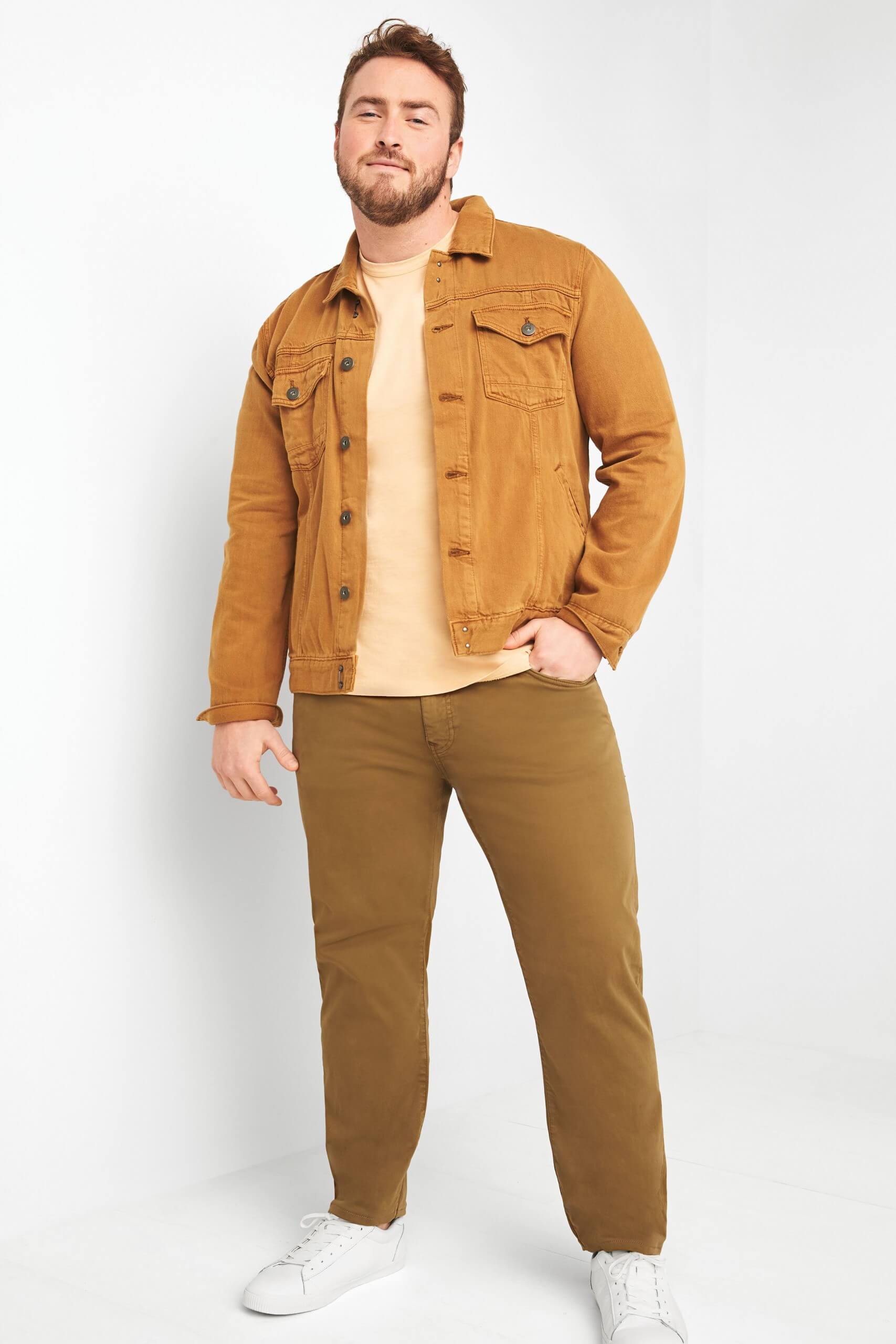 Stitch Fix Men’s model wearing tan denim jacket over yellow t-shirt with brown pants and white sneakers. 