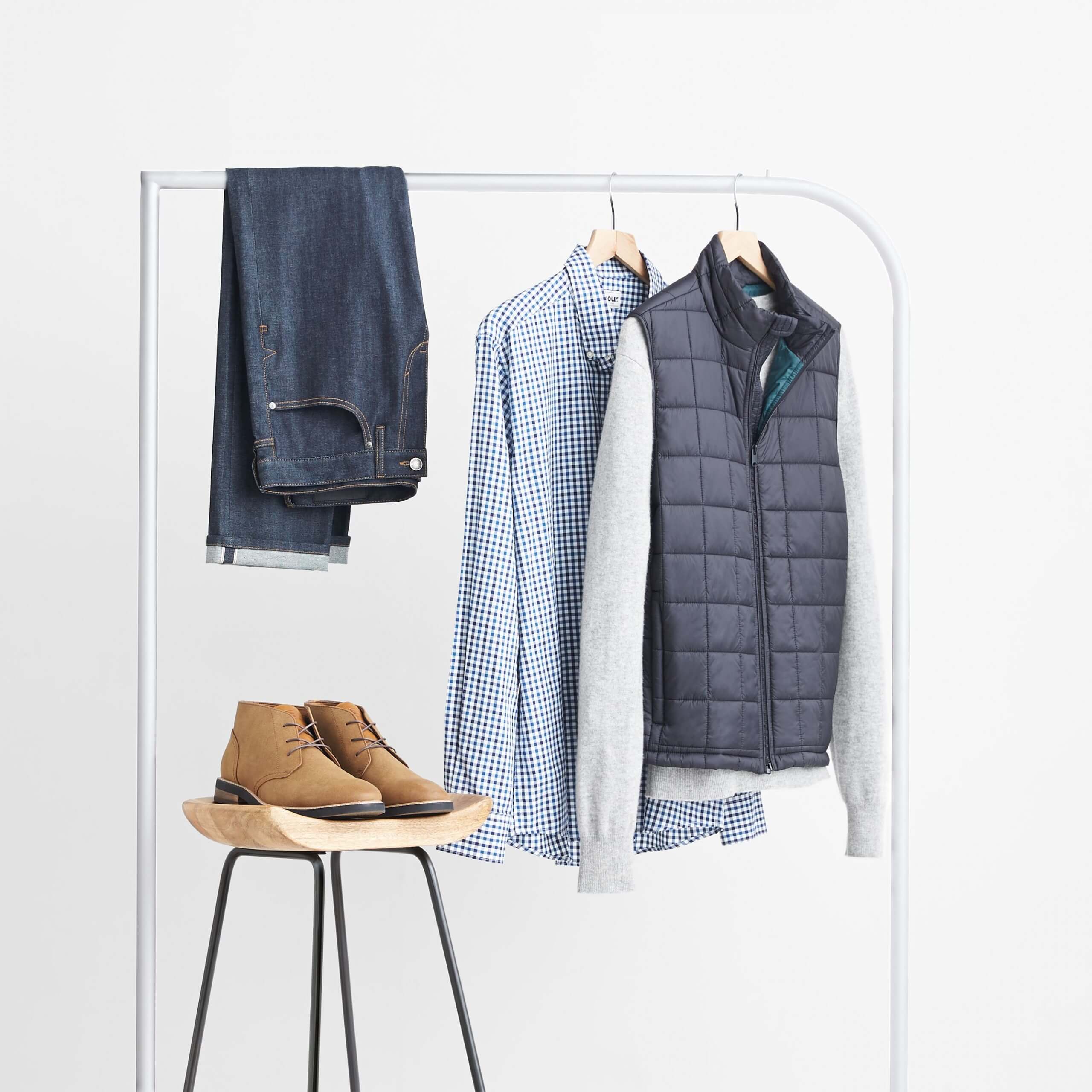 Stitch Fix Men’s dark wash jeans, blue gingham button-down shirt, grey pullover and navy vest hanging on white rack next to brown chukkas on wooden stool.