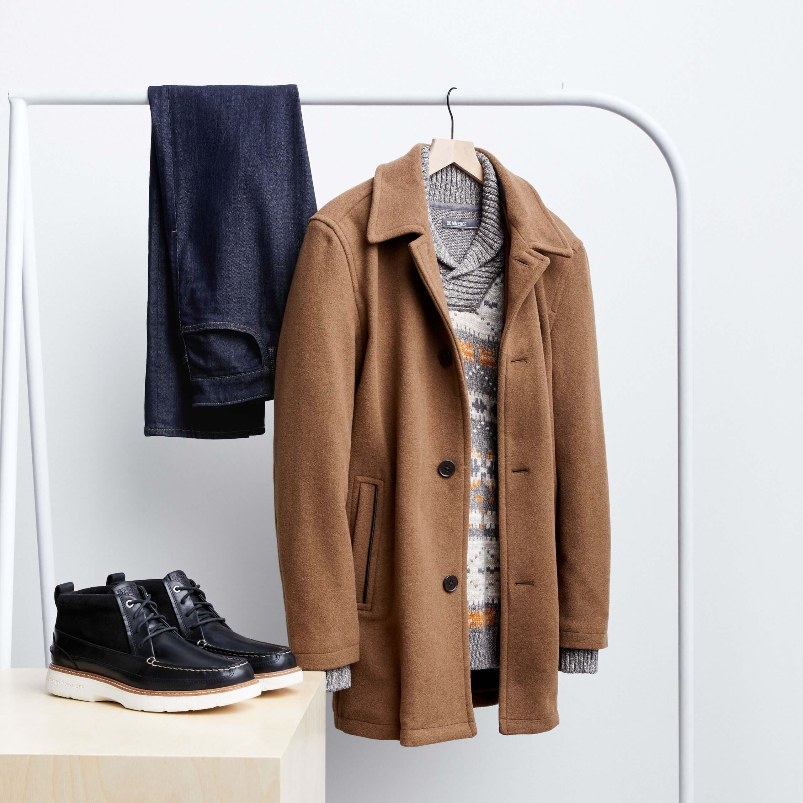 Stitch Fix men's rack image featuring blue jeans folded over rack, brown car coat over grey pullover on hanger, next to black chukka boots on white block.