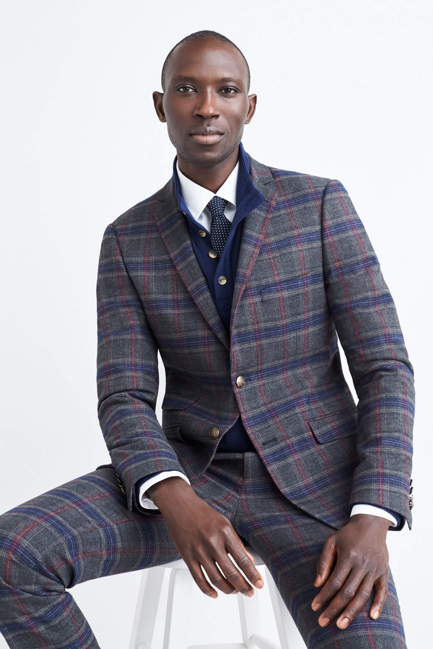 Stitch Fix Men’s model sitting on a white stool wearing a navy plaid suit, white collared shirt and navy blue tie.