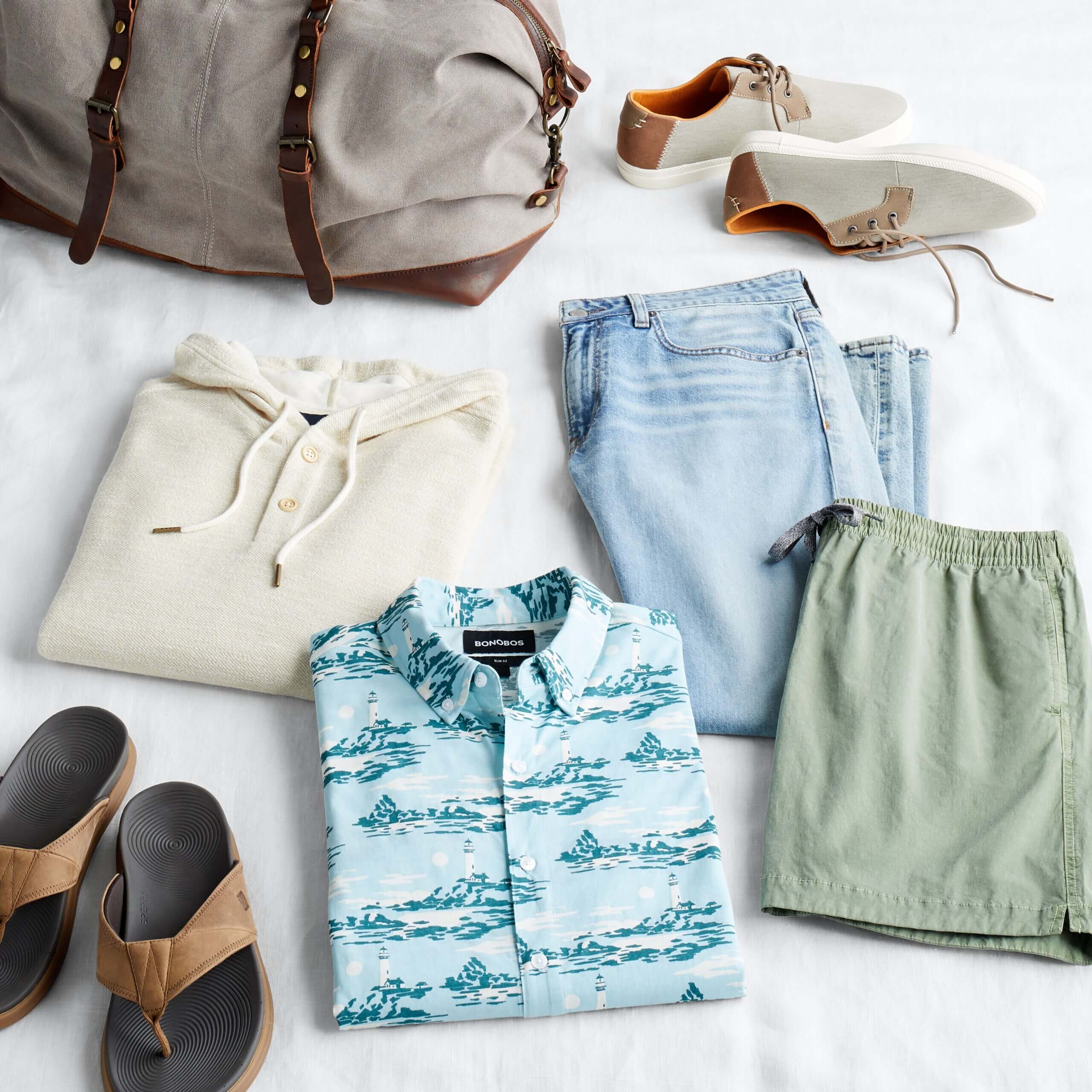 Stitch Fix men's laydown featuring cream hoodie, light wash jeans, green shorts, blue tropical button-down shirt and brown sandals next to duffle bag. 