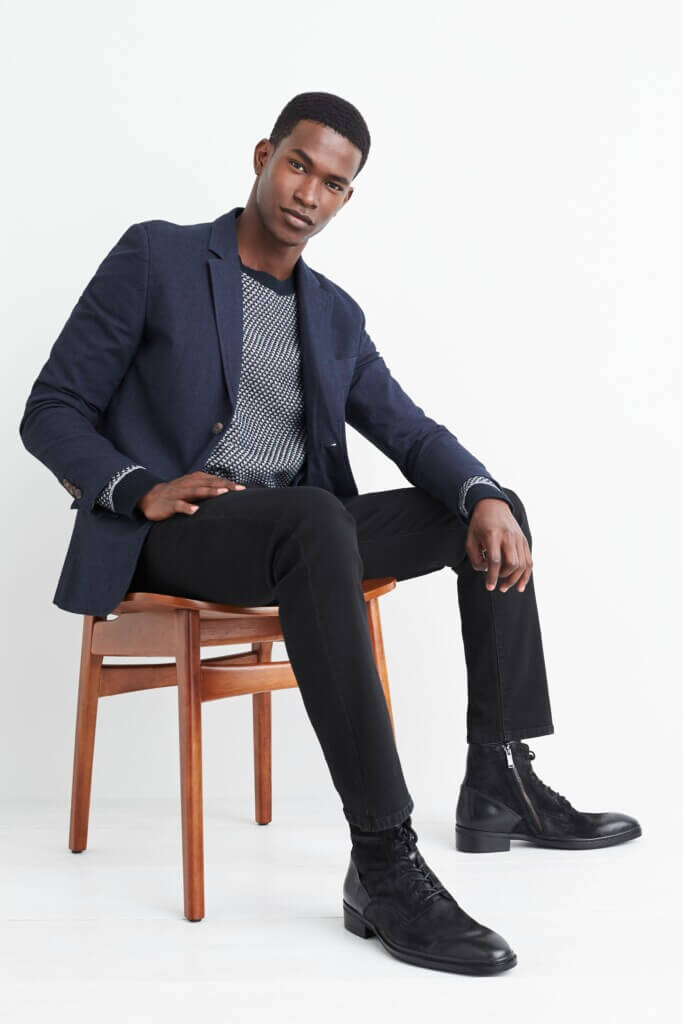 Stitch Fix model wearing men's fashion broad shoulder outfit with navy blazer, patterned navy sweater, black pants and boots.