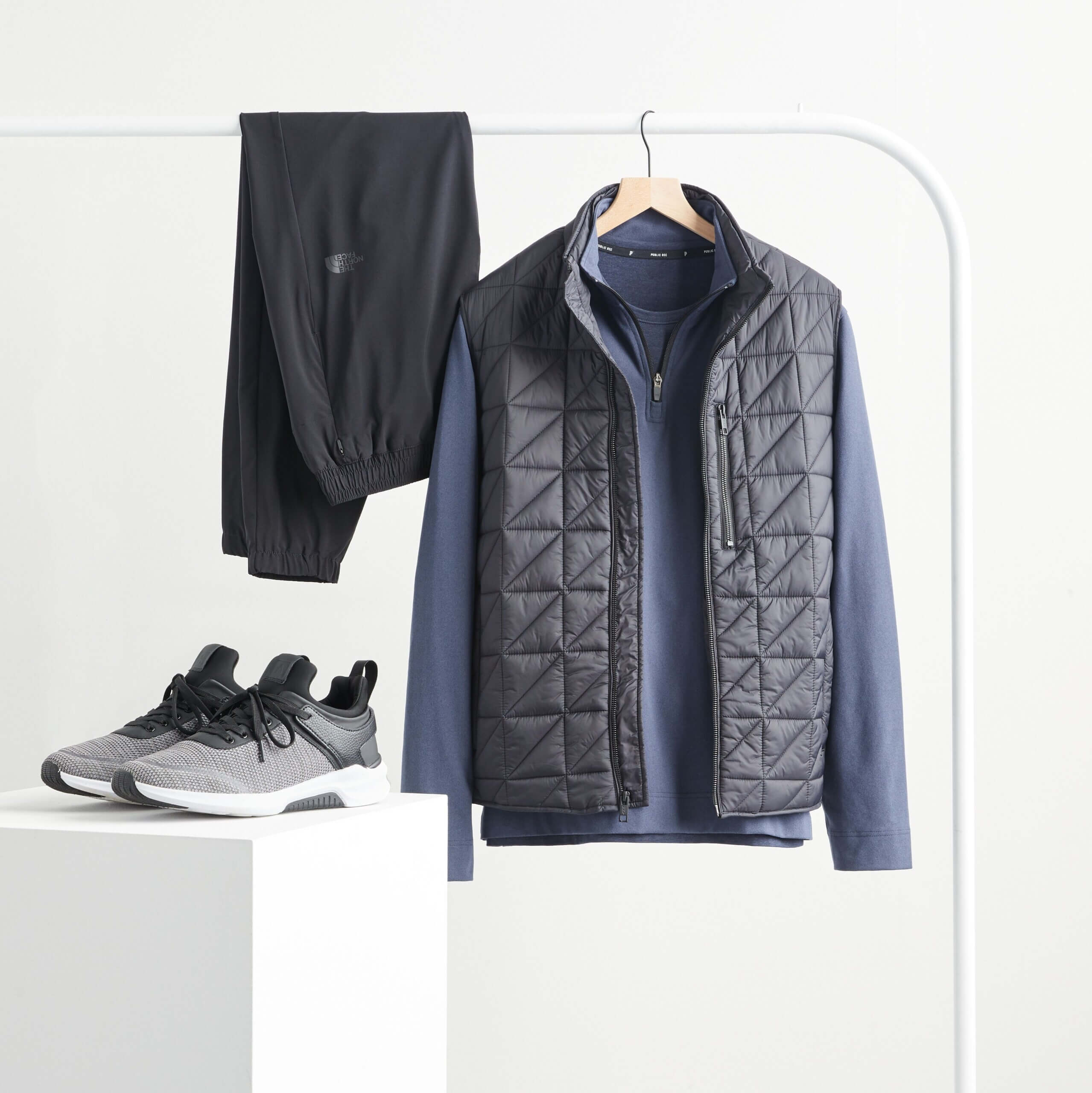 Stitch Fix men's rack image featuring quarter-zip pullover and puffer vest on wooden hanger, black joggers hanging over white rack and performance sneakers on white block.