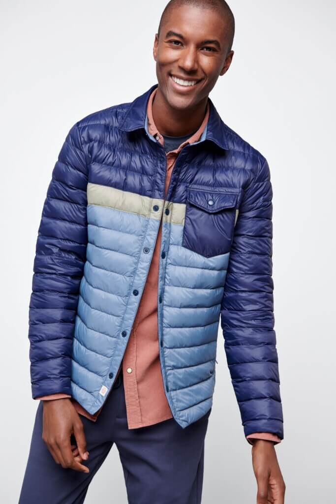 Stitch Fix model wearing men’s broad shoulder outfit with pink button-down shirt, blue color block puffer jacket and navy pants.
