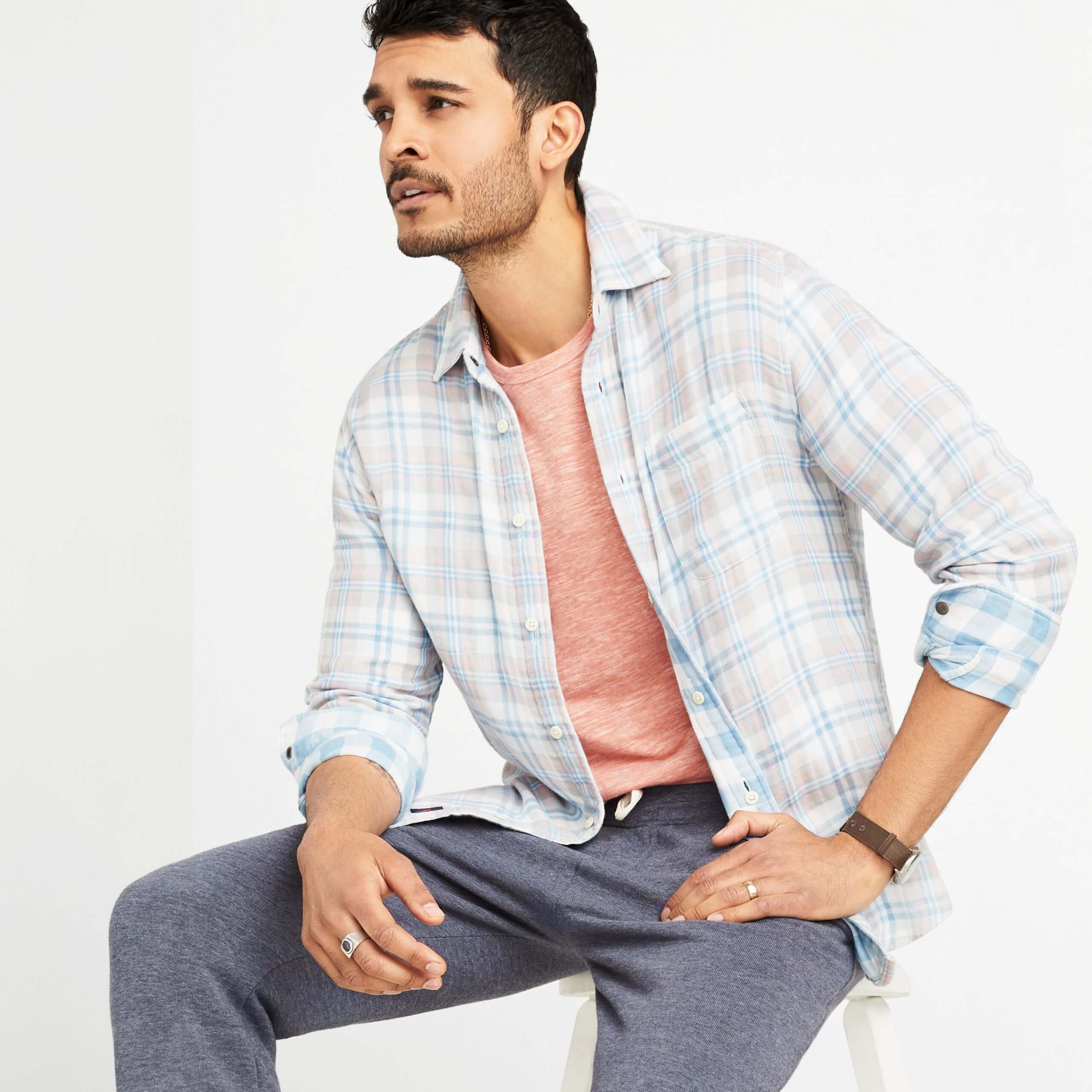 Stitch Fix Men’s model sitting on a white stool wearing pink terry crew sweatshirt under a plaid button-down shirt with fleece pants sitting on white stool.