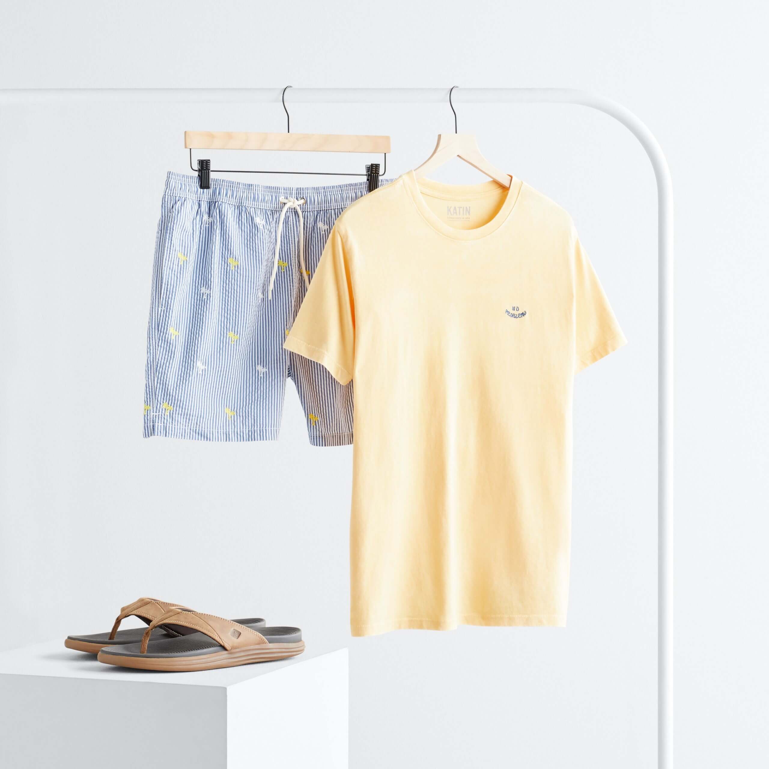 Stitch Fix Men’s rack image with yellow short sleeve graphic tee and blue striped shorts hanging on white rack next to brown flip flops on white block.