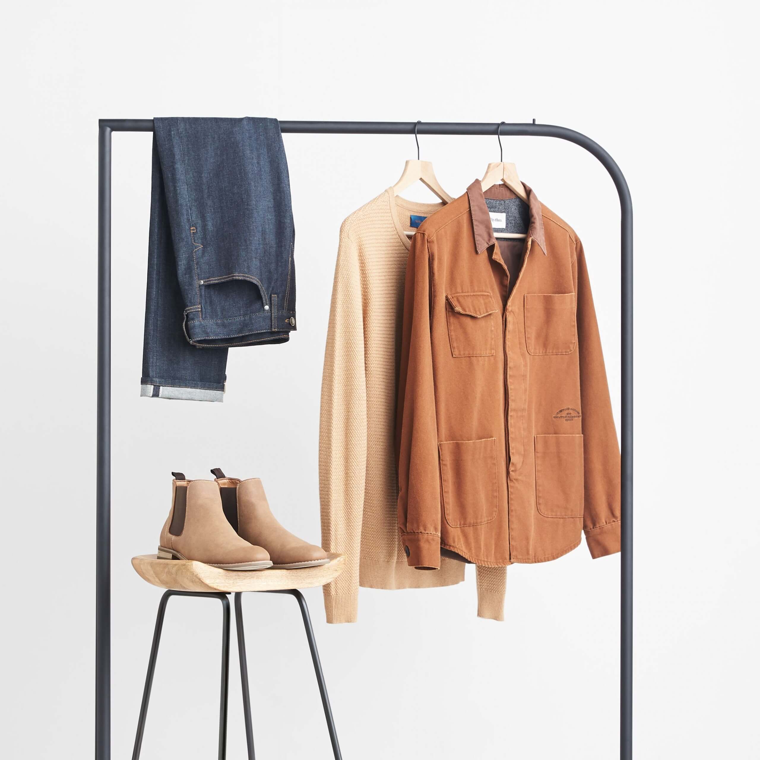 Stitch Fix Men’s brown chore jacket, tan crewneck sweater and slim-fit dark wash jeans hanging on a black rack next to brown boots sitting on a wooden stool. 