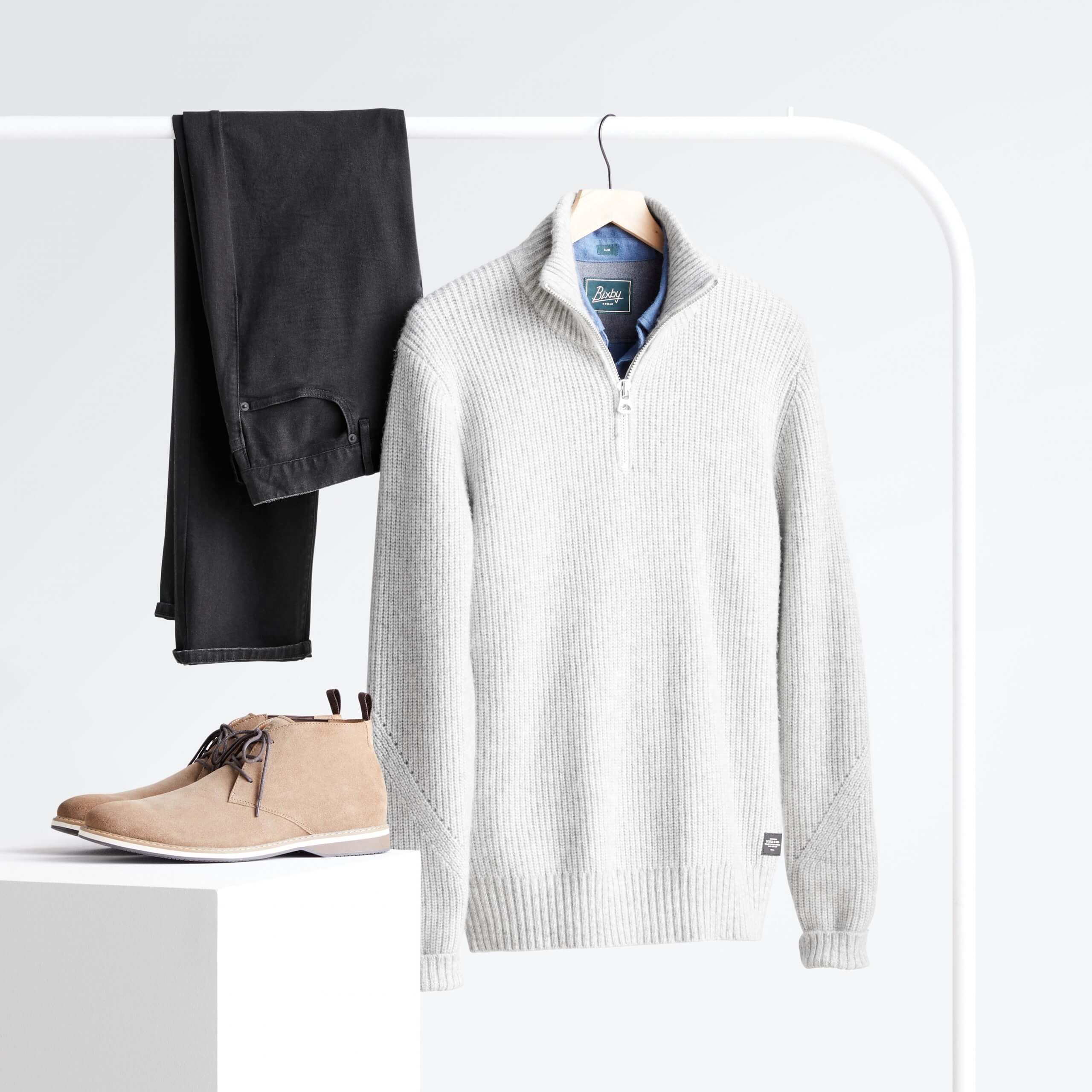 Stitch Fix Men’s light grey quarter zip sweater over blue button-down, black jeans hanging on white rack next to beige chukkas on white podium. 