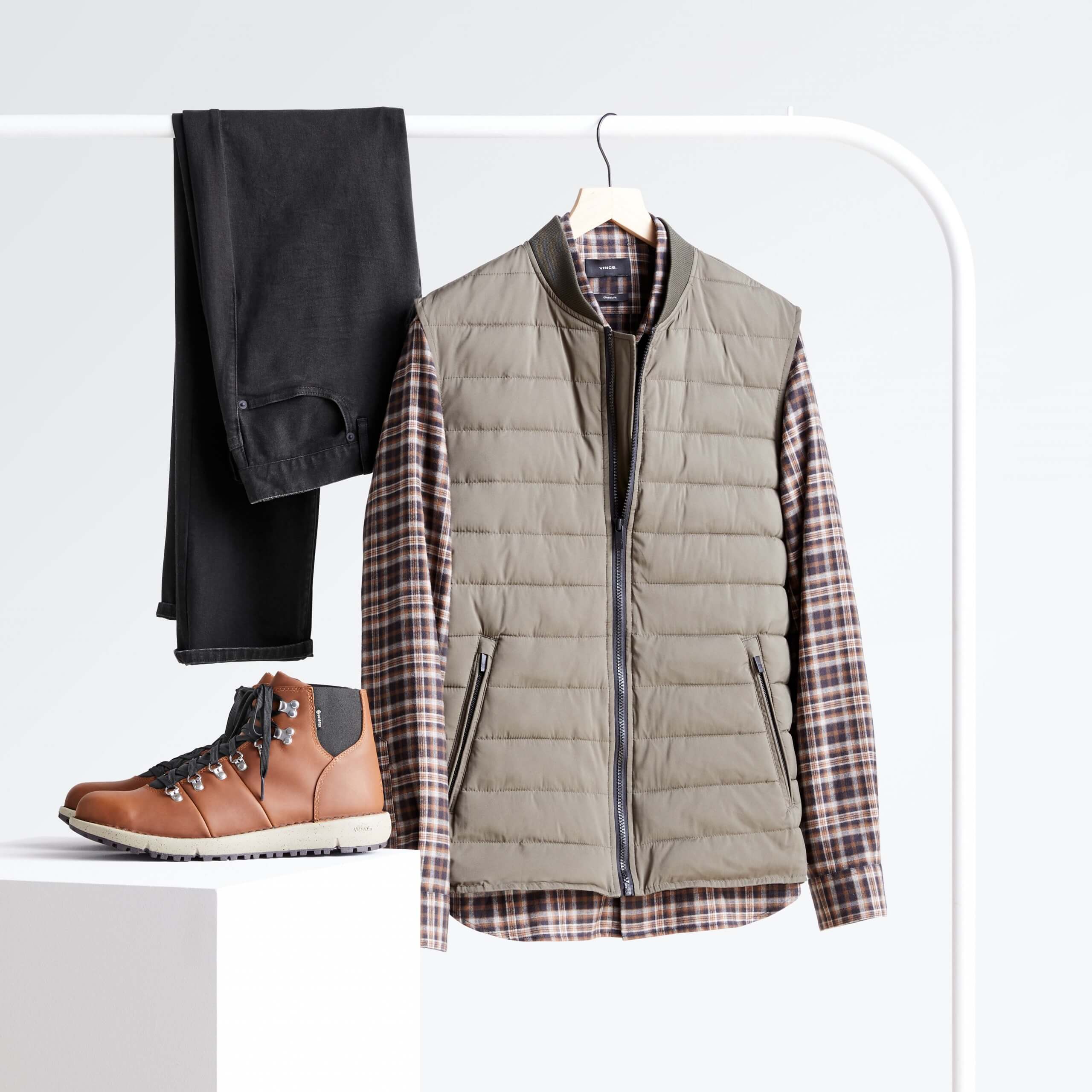 Stitch Fix Men’s grey vest over plaid shirt and black jeans hanging on white rack, next to brown hiker boots on white block. 