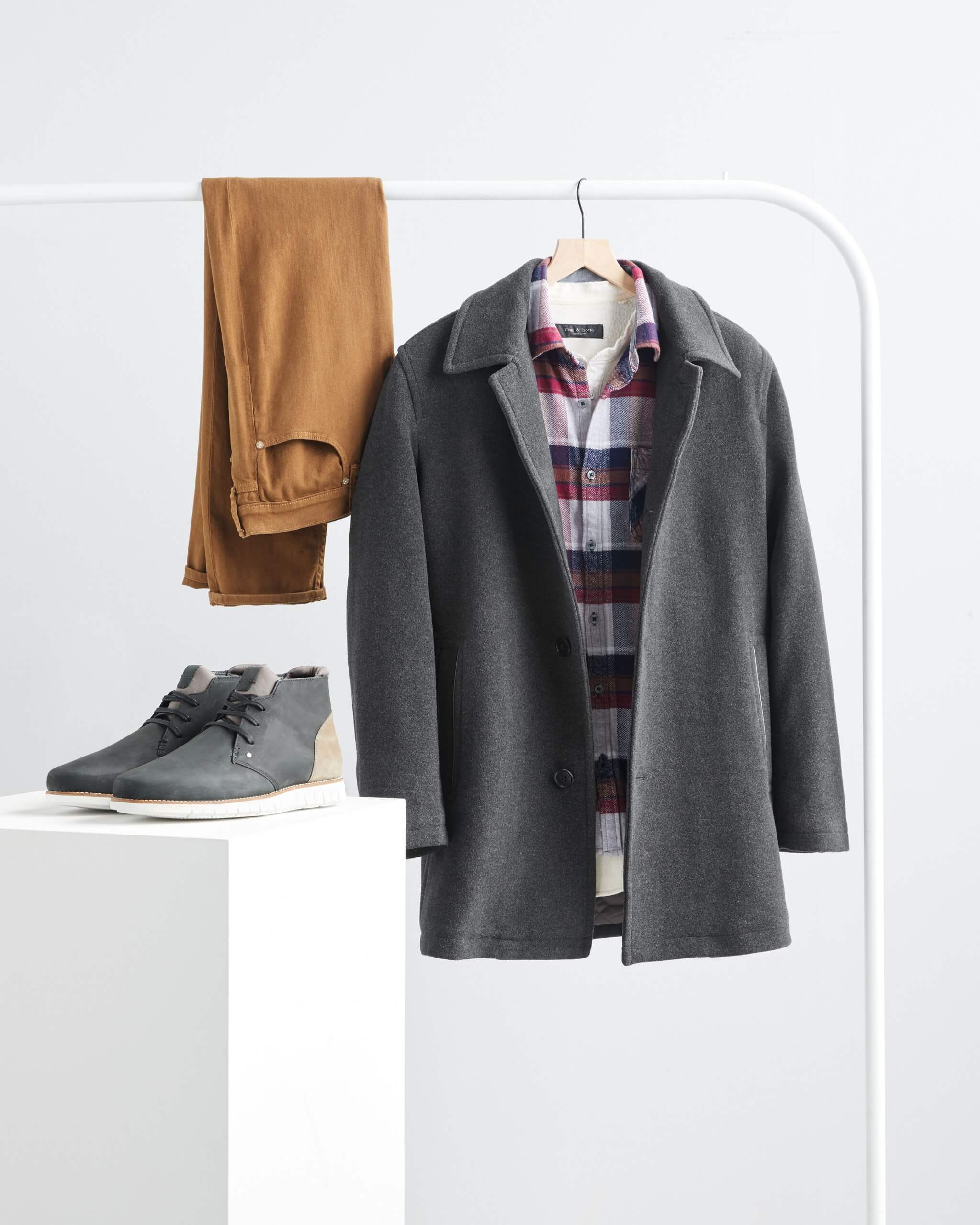 Stitch Fix Men’s grey wool peacoat, red and blue plaid flannel shirt and brown slim-fit jeans hanging on a rack with grey chukkas on a white podium.