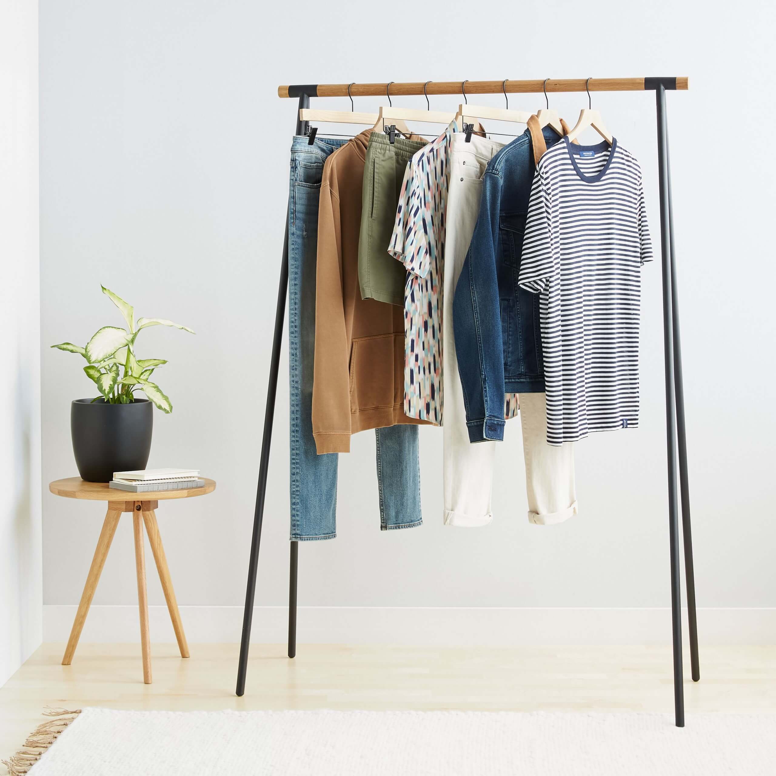 Stitch Fix Men’s blue jeans, tan hoodie, olive shorts, printed T-shirt, white pants, denim jacket and blue T-shirt on a rack next to a wooden table with a plant and two books on top and a rug.