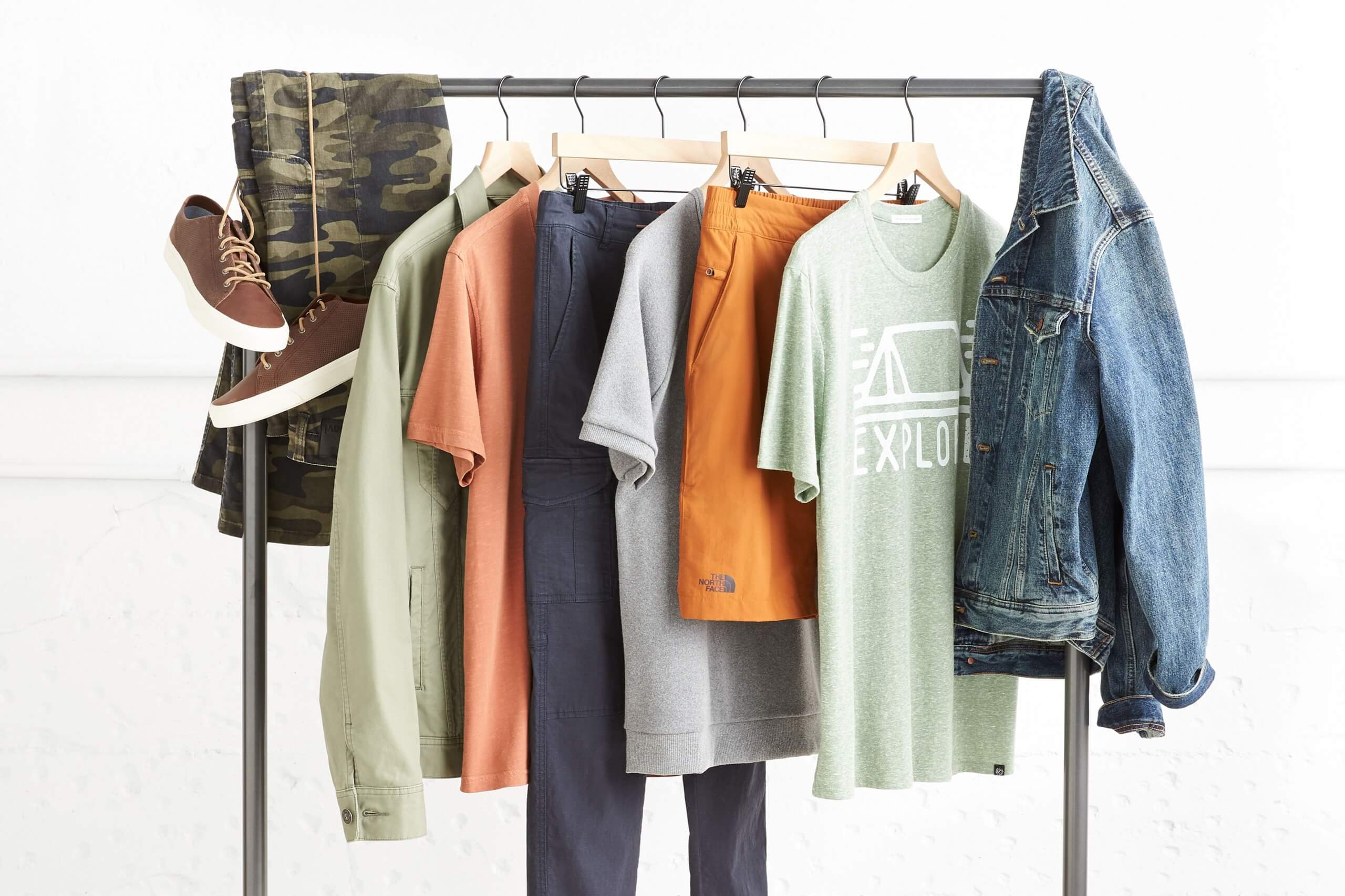 Stitch Fix men’s rack of clothing featuring brown sneakers, camo print shorts, green, orange and blue items and jean jacket.