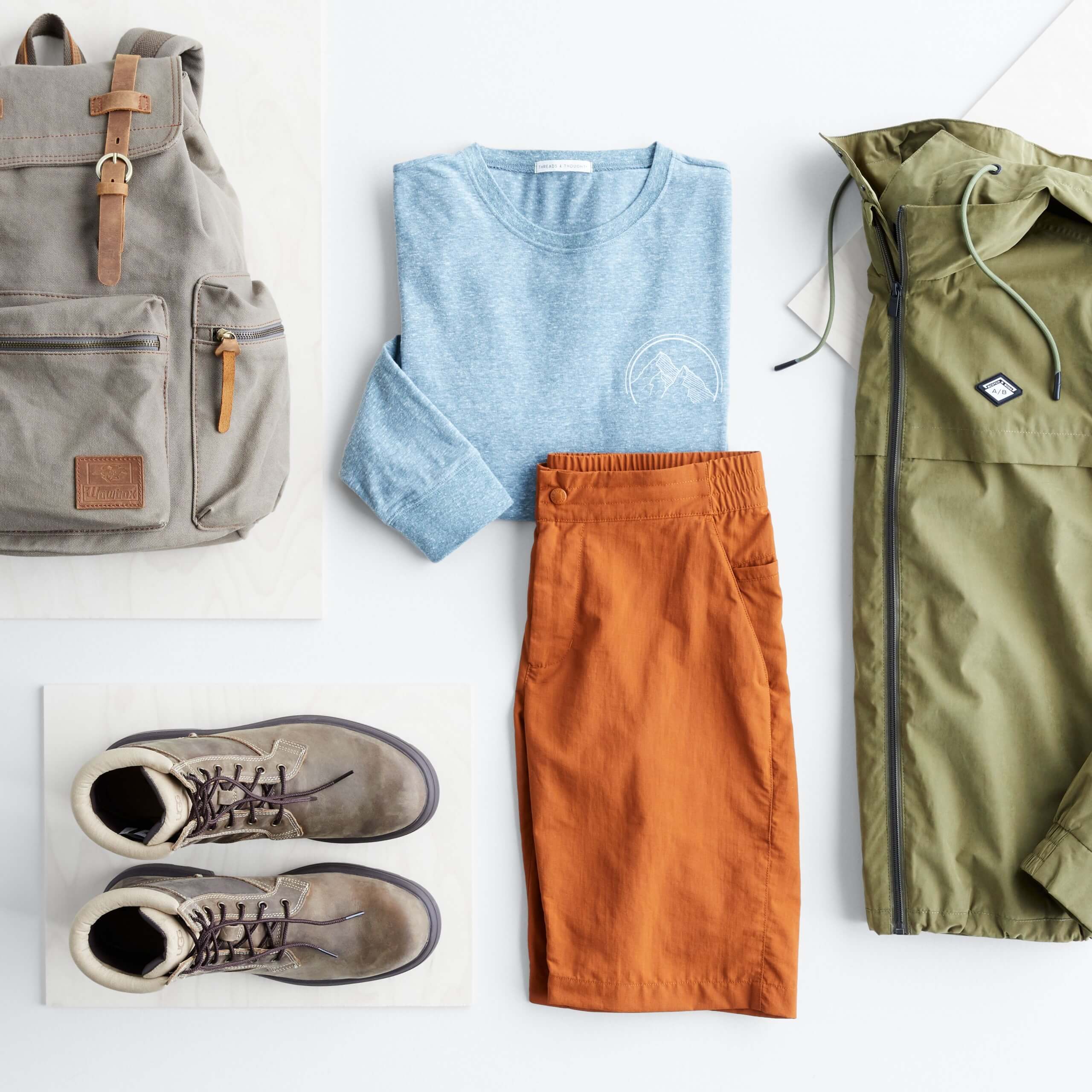Stitch Fix Men’s outfit laydown featuring a blue crewneck, orange shorts, olive jacket, grey boots and grey backpack.