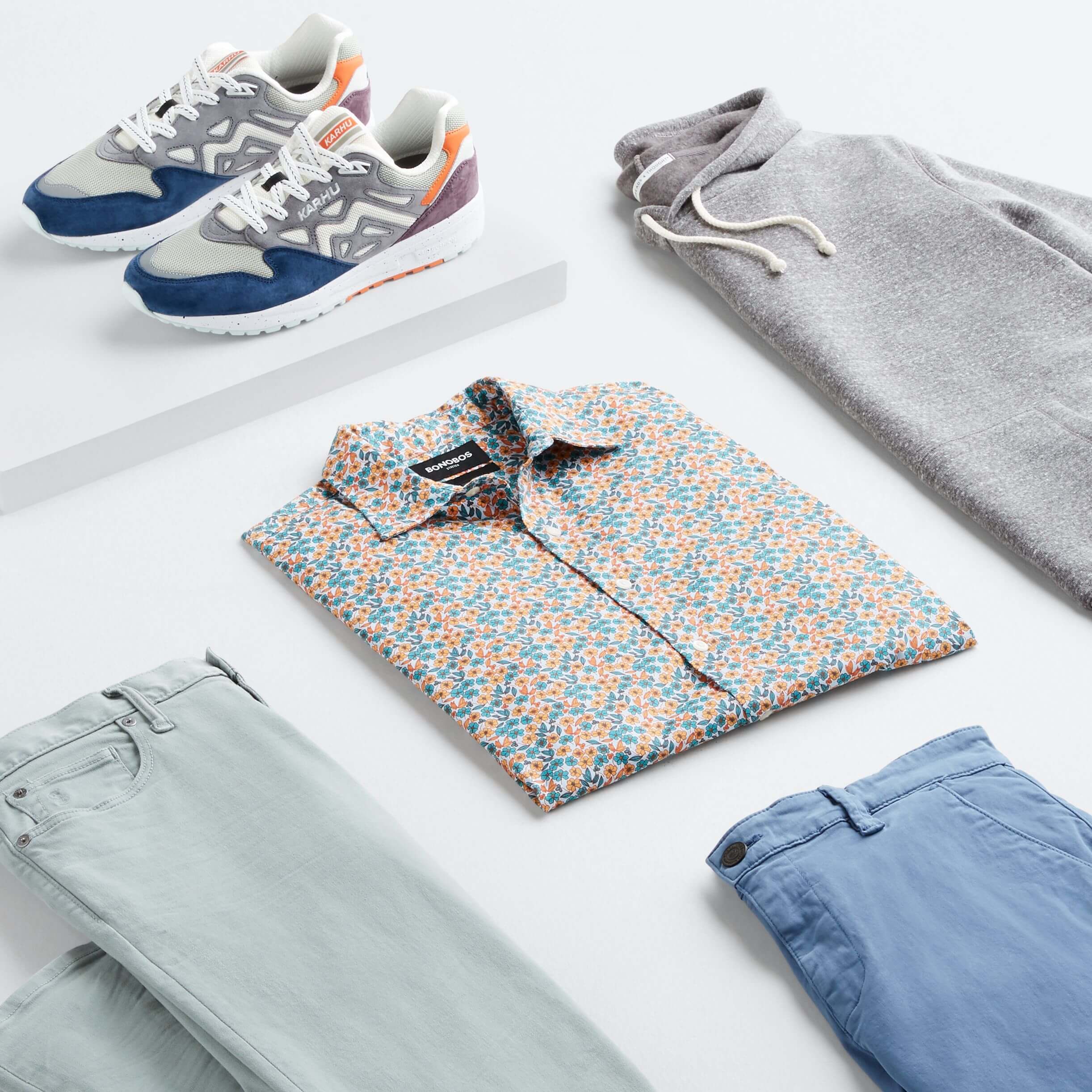 Stitch Fix Men’s outfit laydown featuring an orange and blue printed button-up, light-blue pants, grey hoodie, blue shorts and multi-colored sneakers.