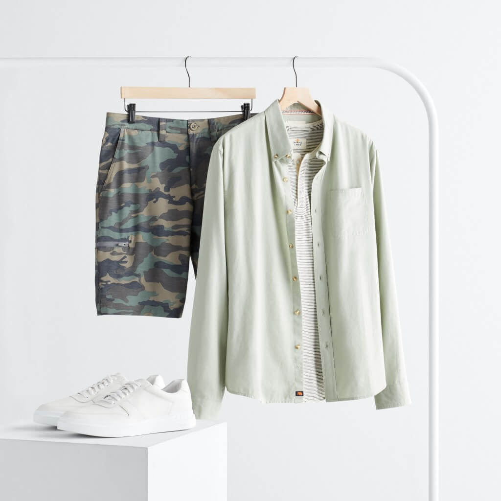 Stitch Fix men’s outfit featuring camo shorts and a light green button-down shirt hanging on a rack and white sneakers on a stand.