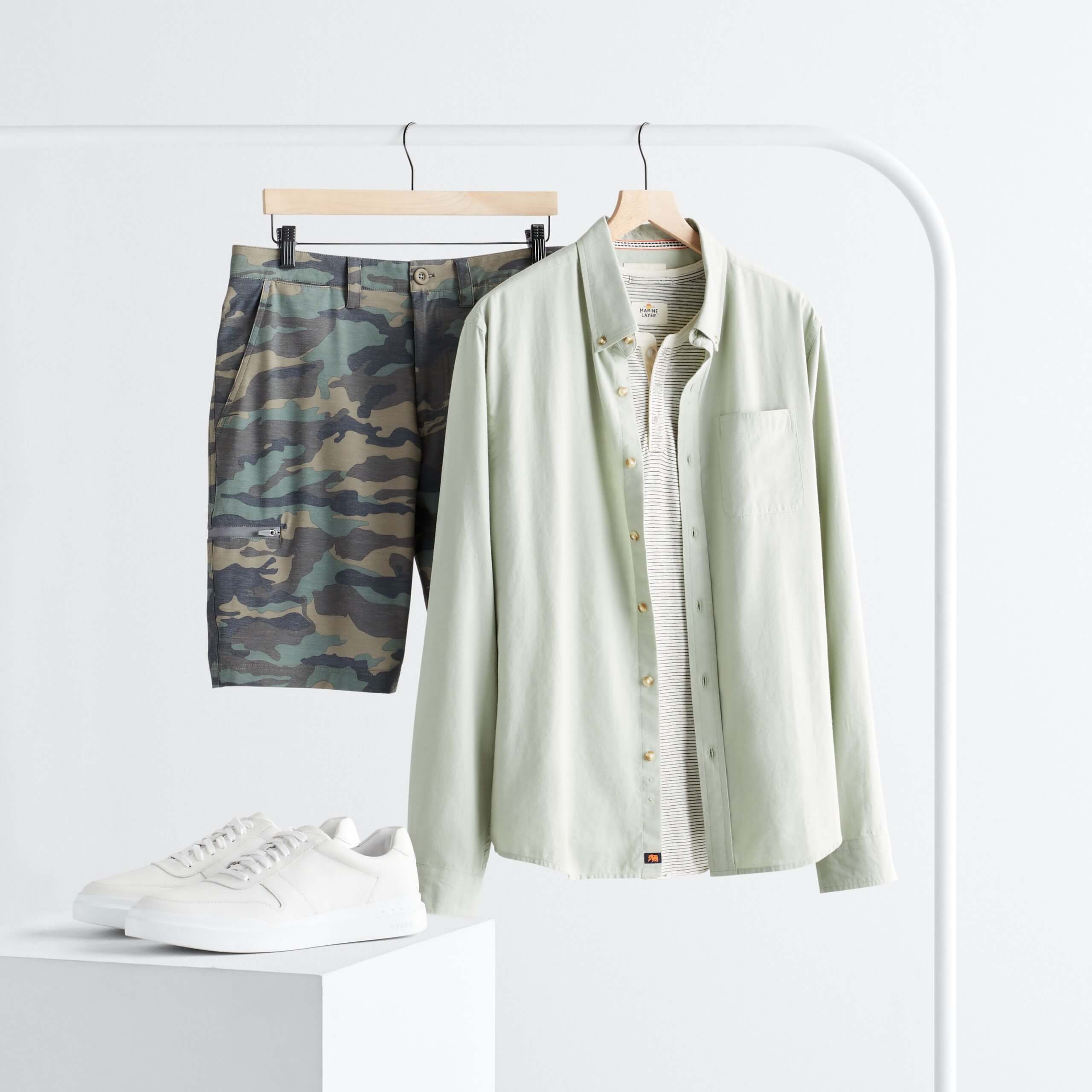 Stitch Fix men’s outfit featuring camo shorts and a light green button-down shirt hanging on a rack and white sneakers on a stand.