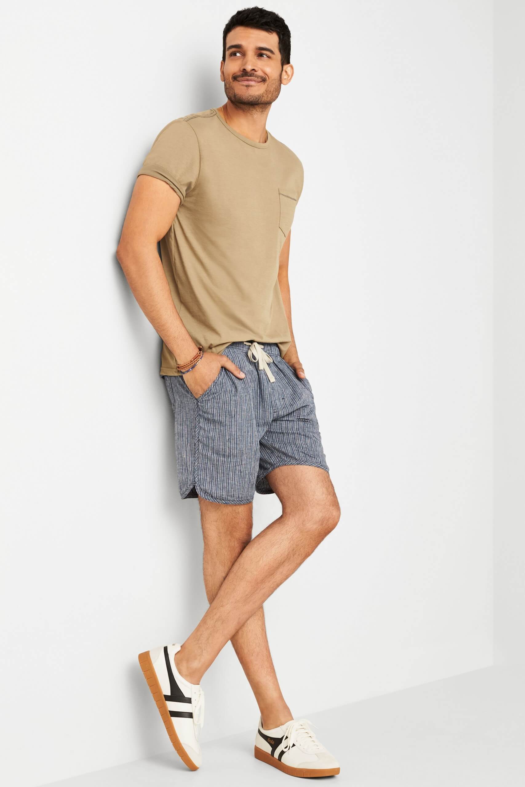 Stitch Fix model wearing men’s tan short-sleeve T-shirt, navy tie-waist shorts with stripes and woven bracelets with sneakers.