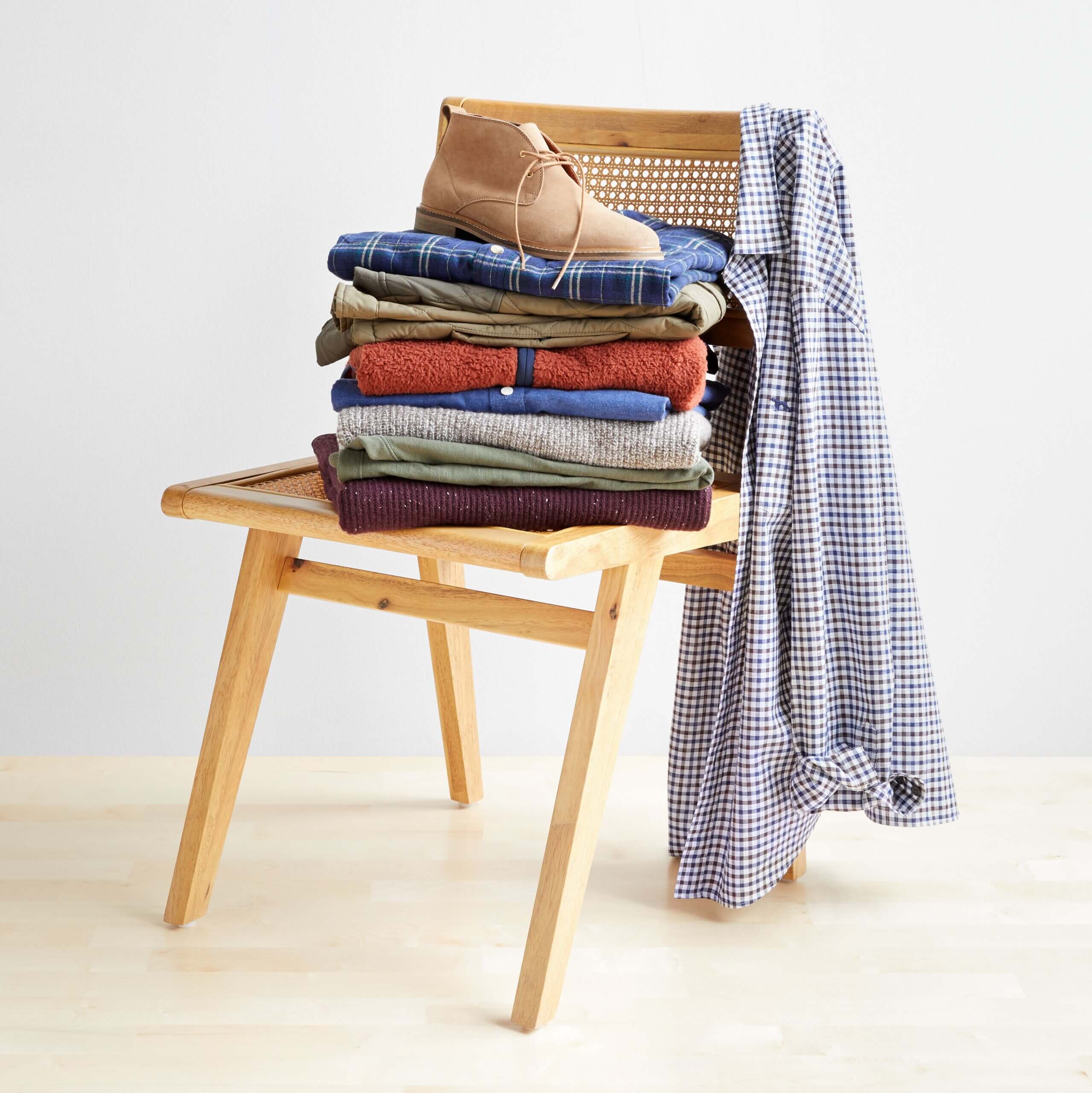 Stitch Fix men’s workleisure collection featuring a selection of sweaters, chinos, jackets, shirts and boots stacked on chair.