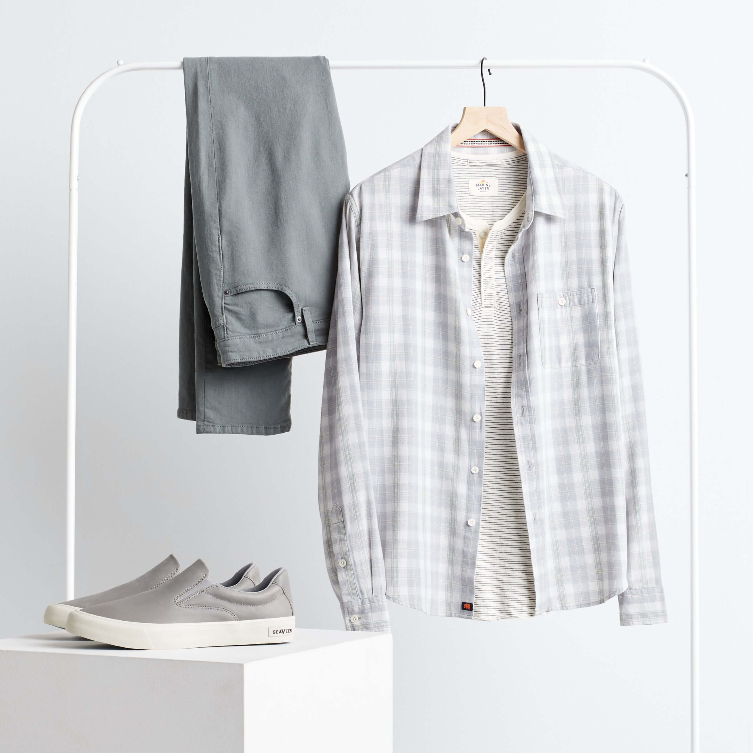 Stitch Fix men’s workleisure outfit with grey chinos draped over clothing rack with striped long-sleeve henley with light grey plaid button-down shirt on hanger and grey slip-on sneakers on platform.