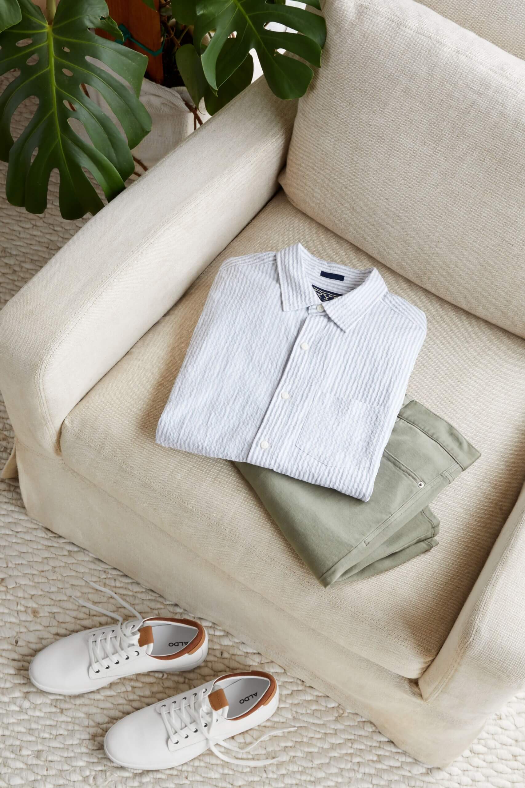 Stitch Fix men’s white button-down and olive chinos folded on top of an off-white chair with a pair of white sneakers on the floor and next to a green plant.