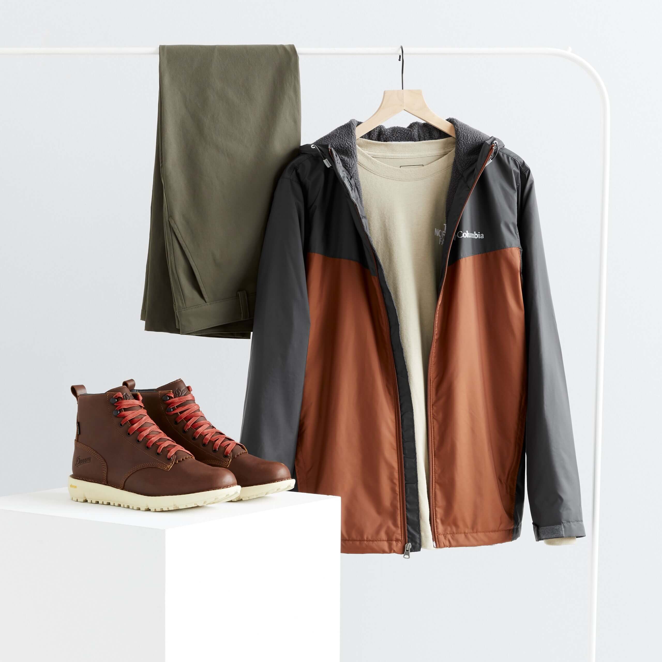 Stitch Fix men’s hiking outfit featuring olive performance pants, beige crew neck T-shirt, orange and grey rain jacket hanging from rack and brown lace-up boots with sport bottom on stand.