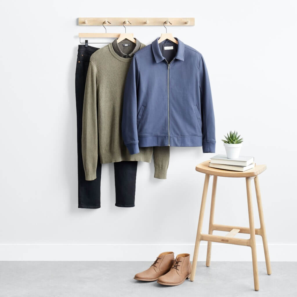 Stitch Fix men’s business casual outfit with jeans, olive knit shirt, blue jacket hanging next to stool with plant and books and taupe boots.