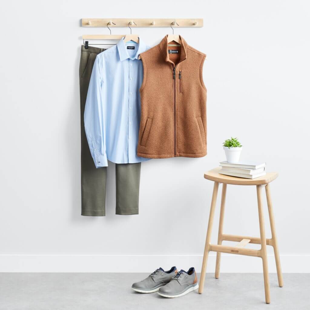 Stitch Fix men’s business casual outfit with blue shirt, burnt orange vest, grey pants hanging next to stool with plant and books and grey shoes.