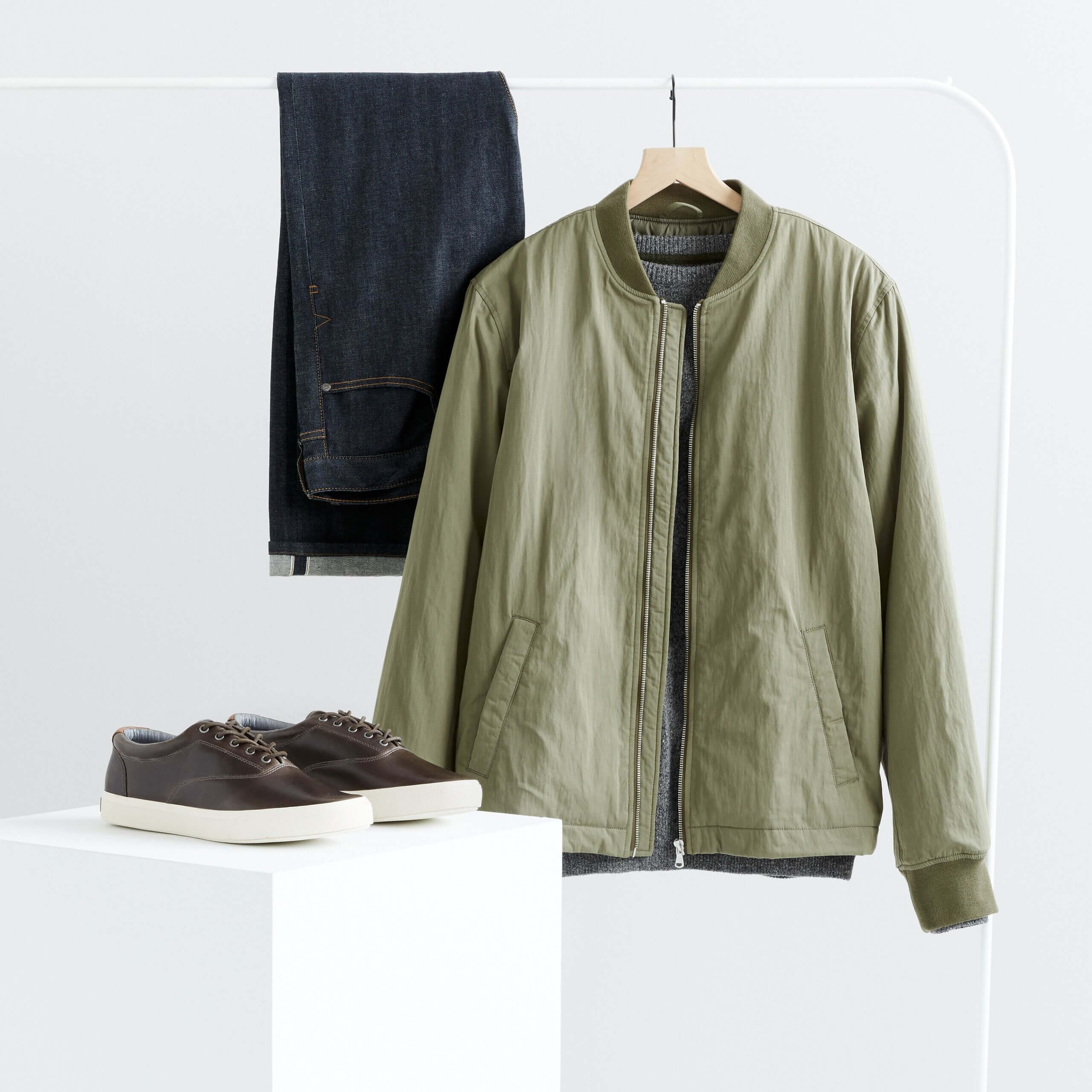 Stitch Fix men’s workleisure outfit featuring blue jeans and olive green bomber jacket on clothing rack with brown leather sneakers on pedestal.
