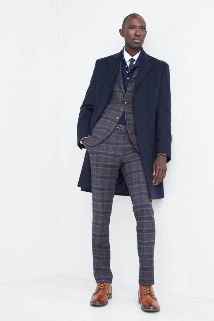 Stitch Fix men's New Year's Eve outfit on model featuring a navy coat over grey, navy and burgundy plaid suit, navy sweater, white button-down, navy tie and brown dress shoes.