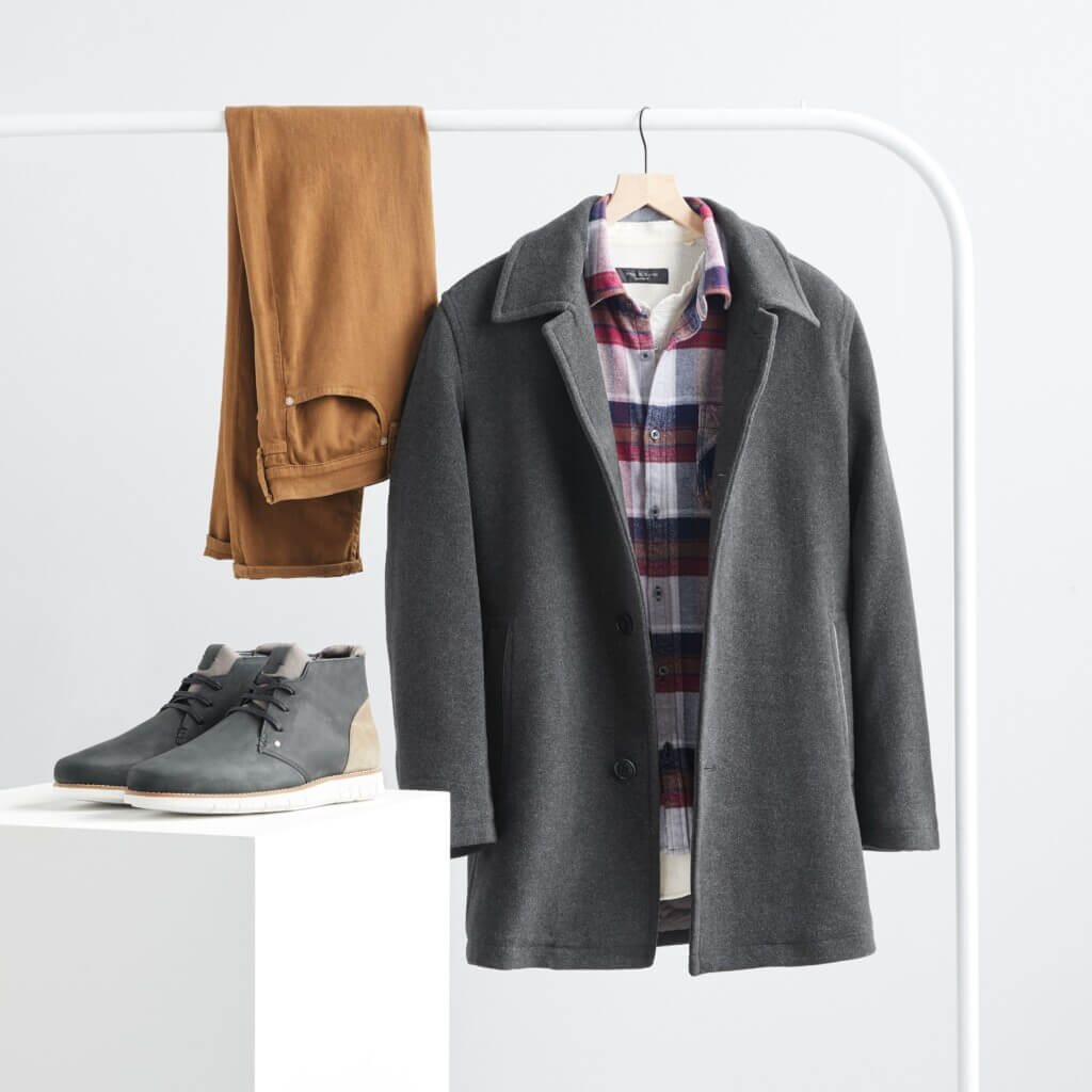 Stitch Fix men’s Christmas outfit featuring gray coat over white, burgundy and navy button-down hanging on rack next to brown jeans and gray chukka boots.