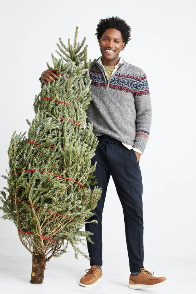 Stitch Fix men’s Christmas outfit on model holding Christmas tree featuring gray, navy and burgundy Fair Isle sweater over white collared shirt and yellow tee, navy jogger pants and brown sneakers.