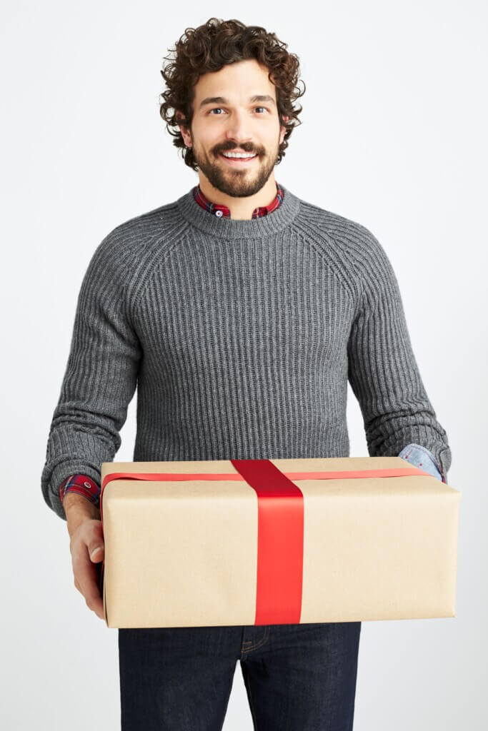 Stitch Fix men's Christmas outfit on model holding a present featuring grey sweater over red collared shirt and blue jeans.
