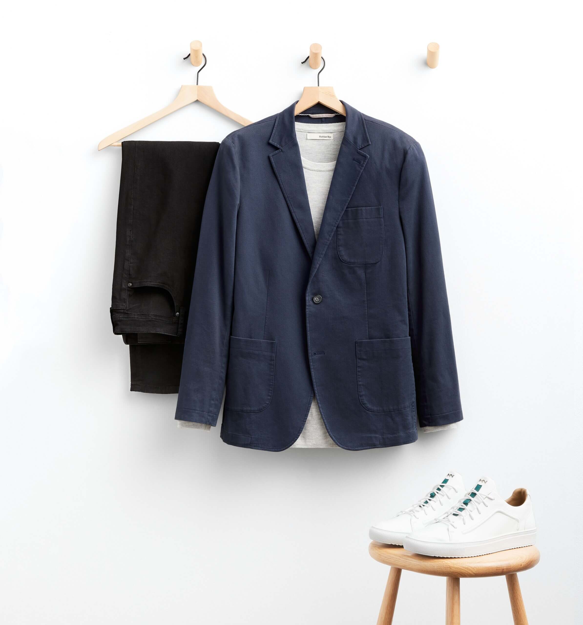 Stitch Fix men’s outfit for shorter men featuring black pants, white shirt and navy blazer hanging next to white court shoes sitting on wood stool.