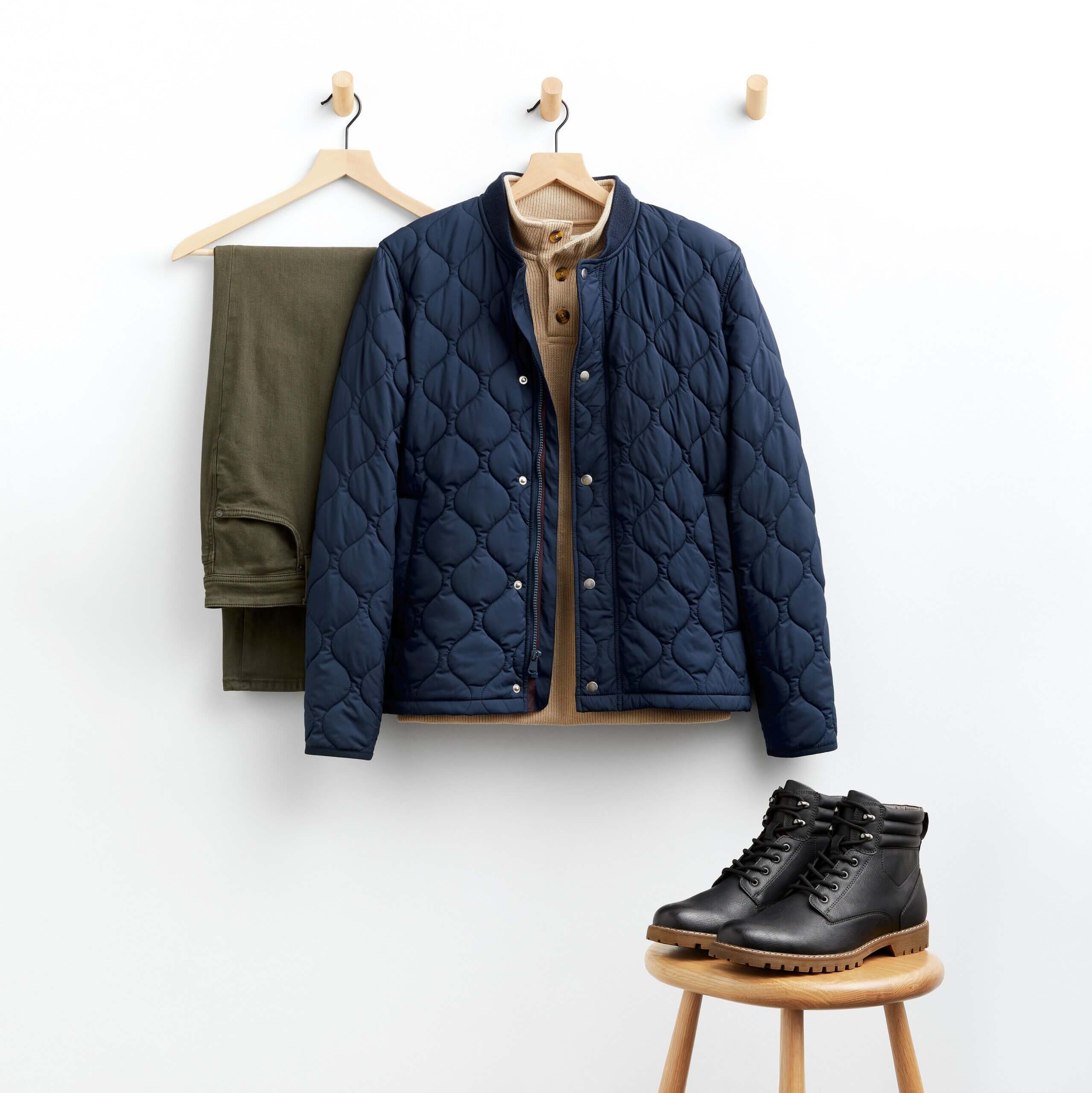Stitch Fix men’s outfit for shorter men featuring green pants, tan sweater and navy quilted puffer jacket hanging next to black hiking boots sitting on wooden stool.