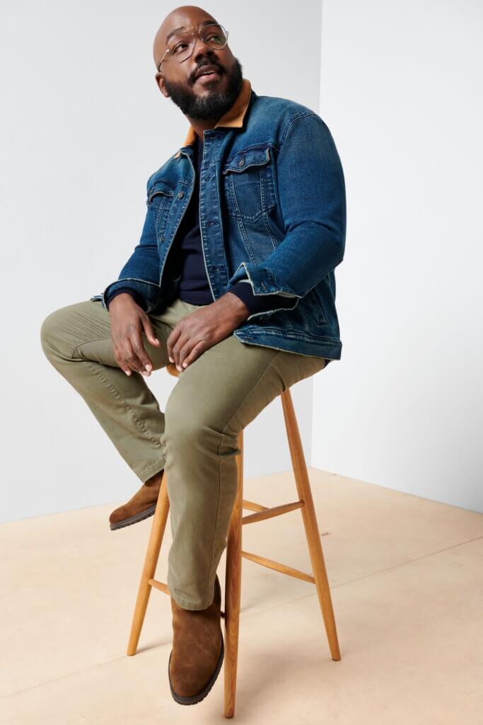 Stitch Fix men’s model sitting on stool wearing outfit featuring blue denim jacket, olive green jeans and brown boots.