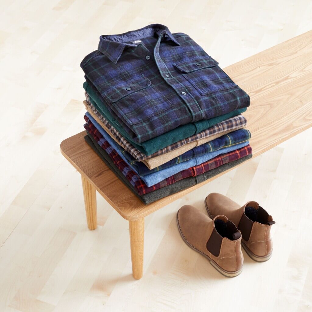 Stitch Fix men’s button-up flannel shirts in various colors and prints folded on a wooden bench next to brown Chelsea boots.