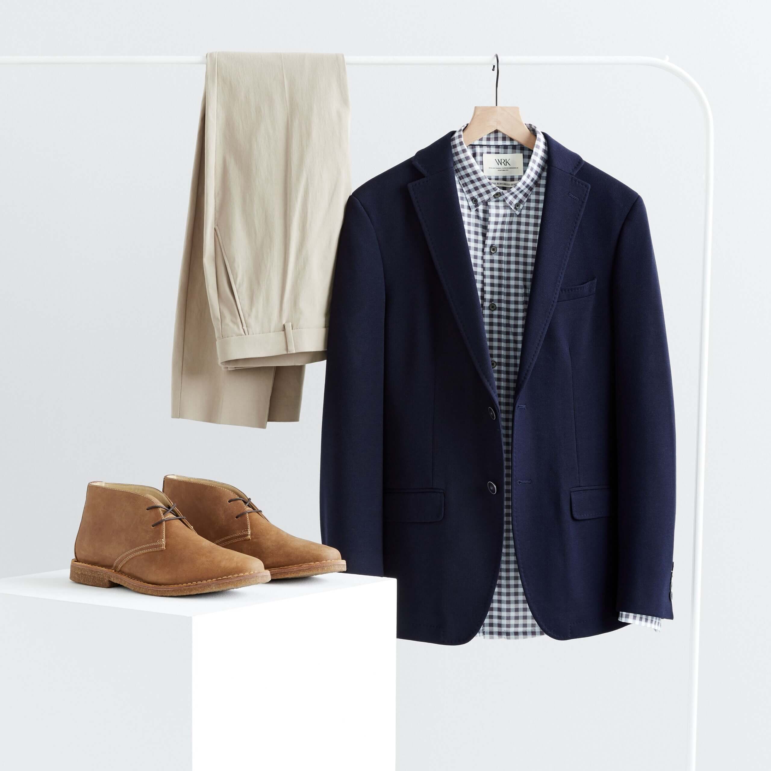 Stitch Fix men's spring wedding guest attire featuring khaki pants, gingham button-down shirt, blue blazer on clothing rack and brown chukkas on platform..